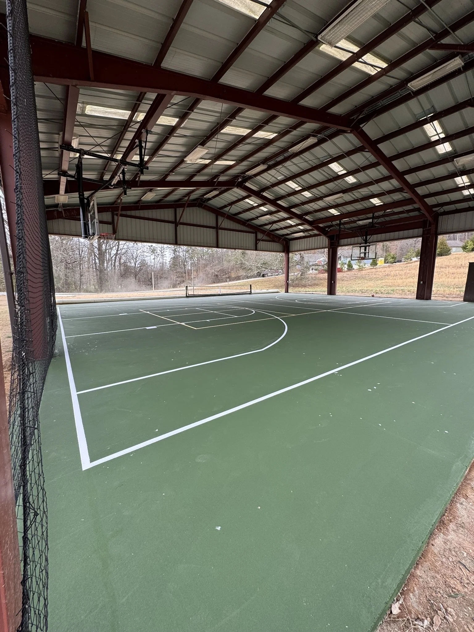 Indoor tennis or basketball court with green flooring, white lines, and a net, under a metal arched roof with skylights, surrounded by a mesh fence and outdoor scenery visible through openings.