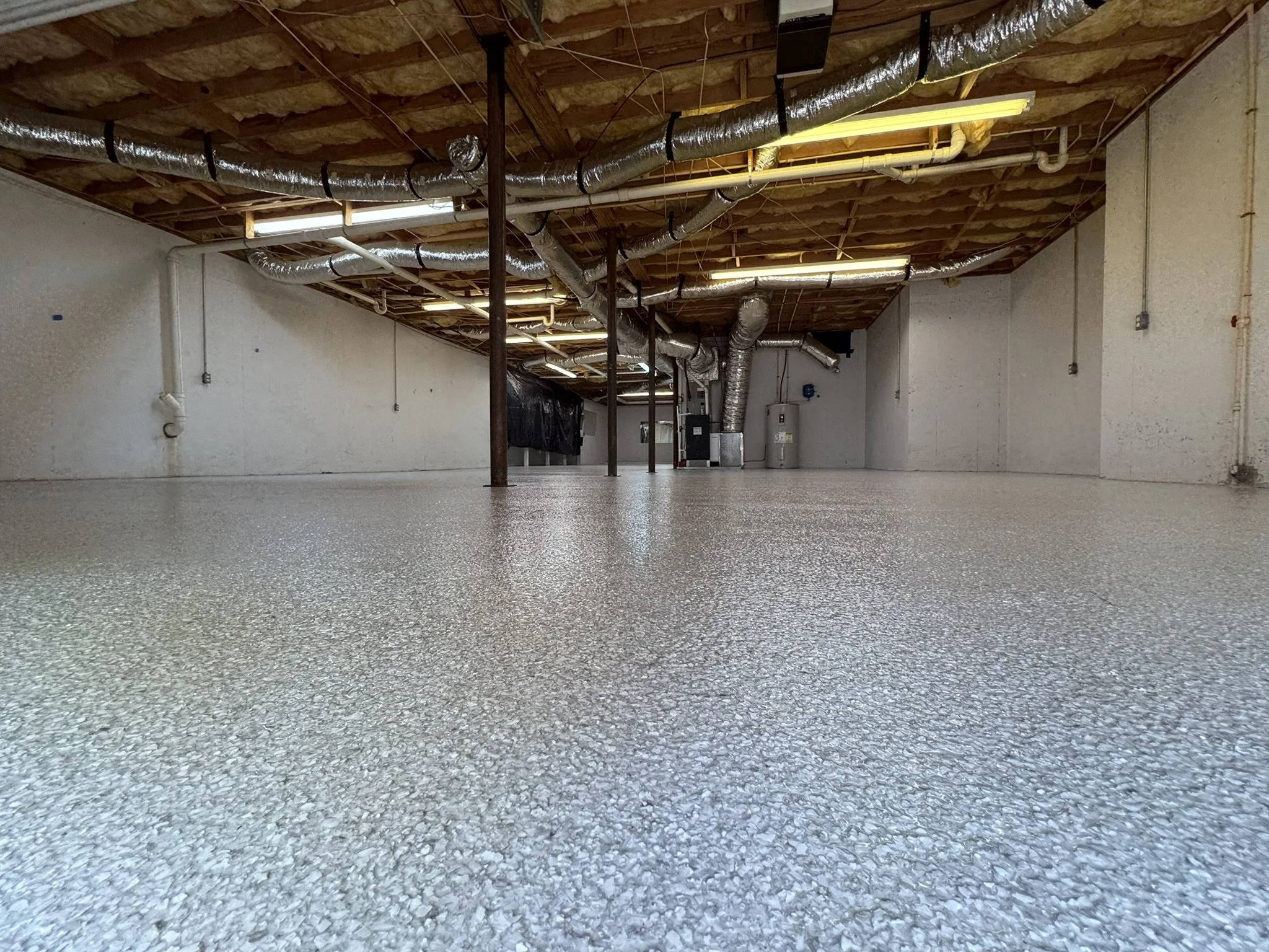 Empty basement with newly painted epoxy floor, exposed ductwork, wooden ceiling, and plumbing pipes.