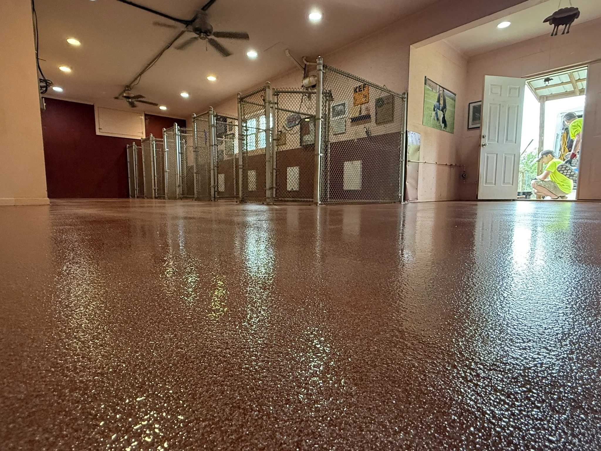 Empty indoor space with polished, shiny reddish-brown floor, chain-link fenced partitions, posters on the wall, and open door showing workers outside.