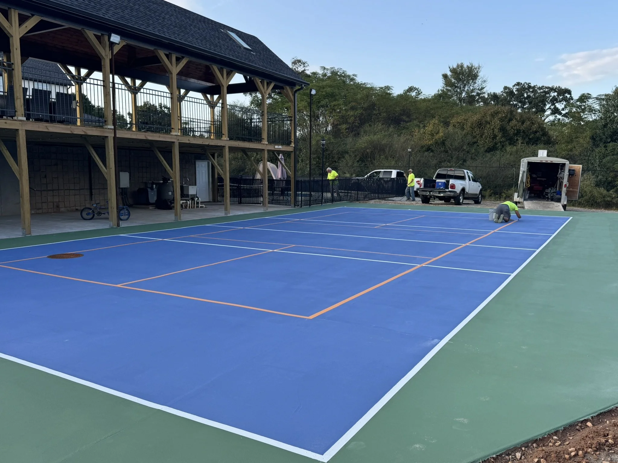 Workers installing a new blue tennis or pickleball court with white and orange boundary lines outside near a two-story wooden building with a balcony.
