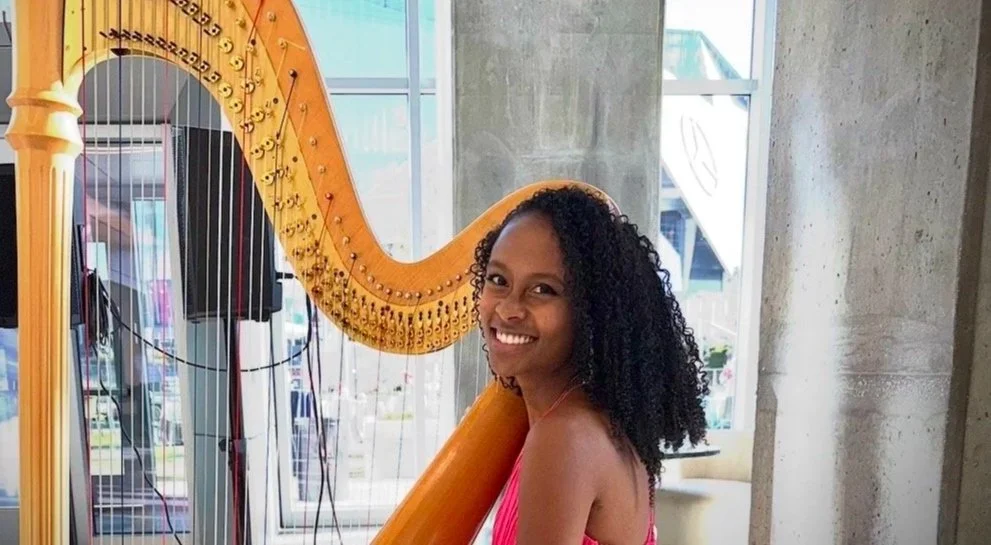 A woman with curly black hair smiling and posing next to a large gold harp.