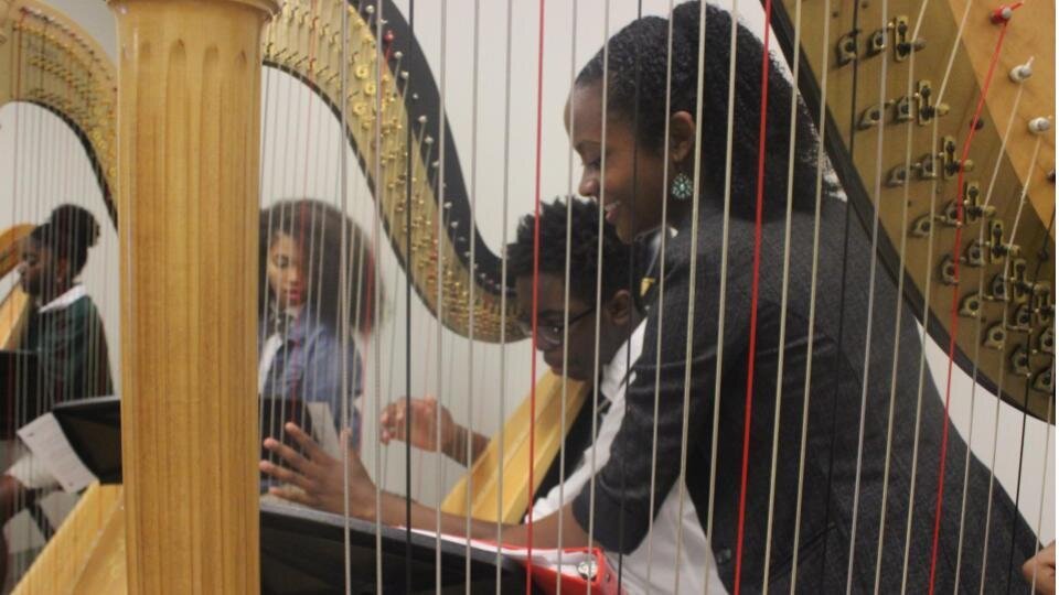 Angelica teaches a group of high school harp students. She is wearing a grey blazer and teaching a young male harp student wearing a school uniform.