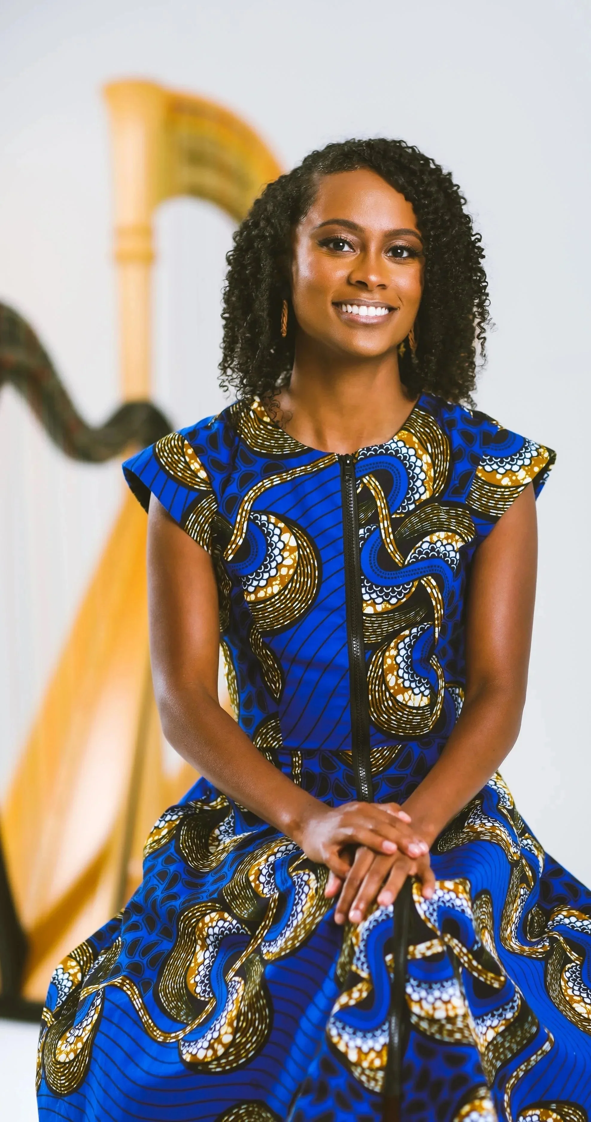Harpist Angelica Hairston sitting wearing a colorful dress with African print in front of harps.