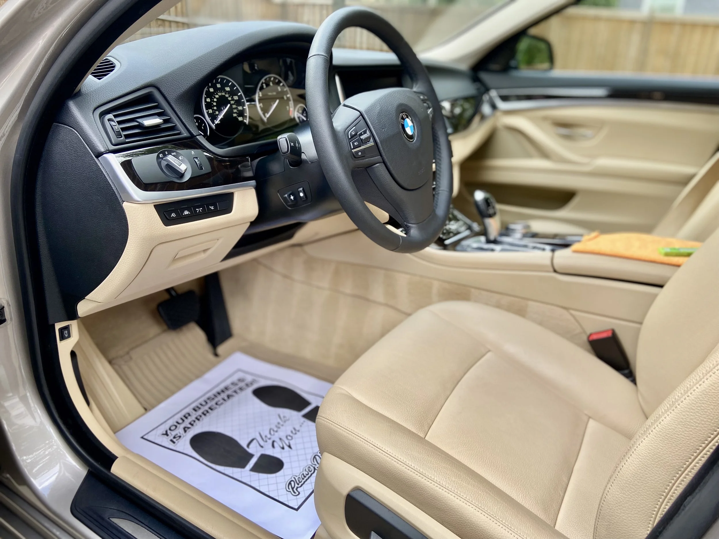 The interior of a BMW car showing the steering wheel, dashboard with gauges, and beige leather seats with auto detailing brush lines in the carpet, There is a paper floor mat with a footprint design and some text on the driver's side.