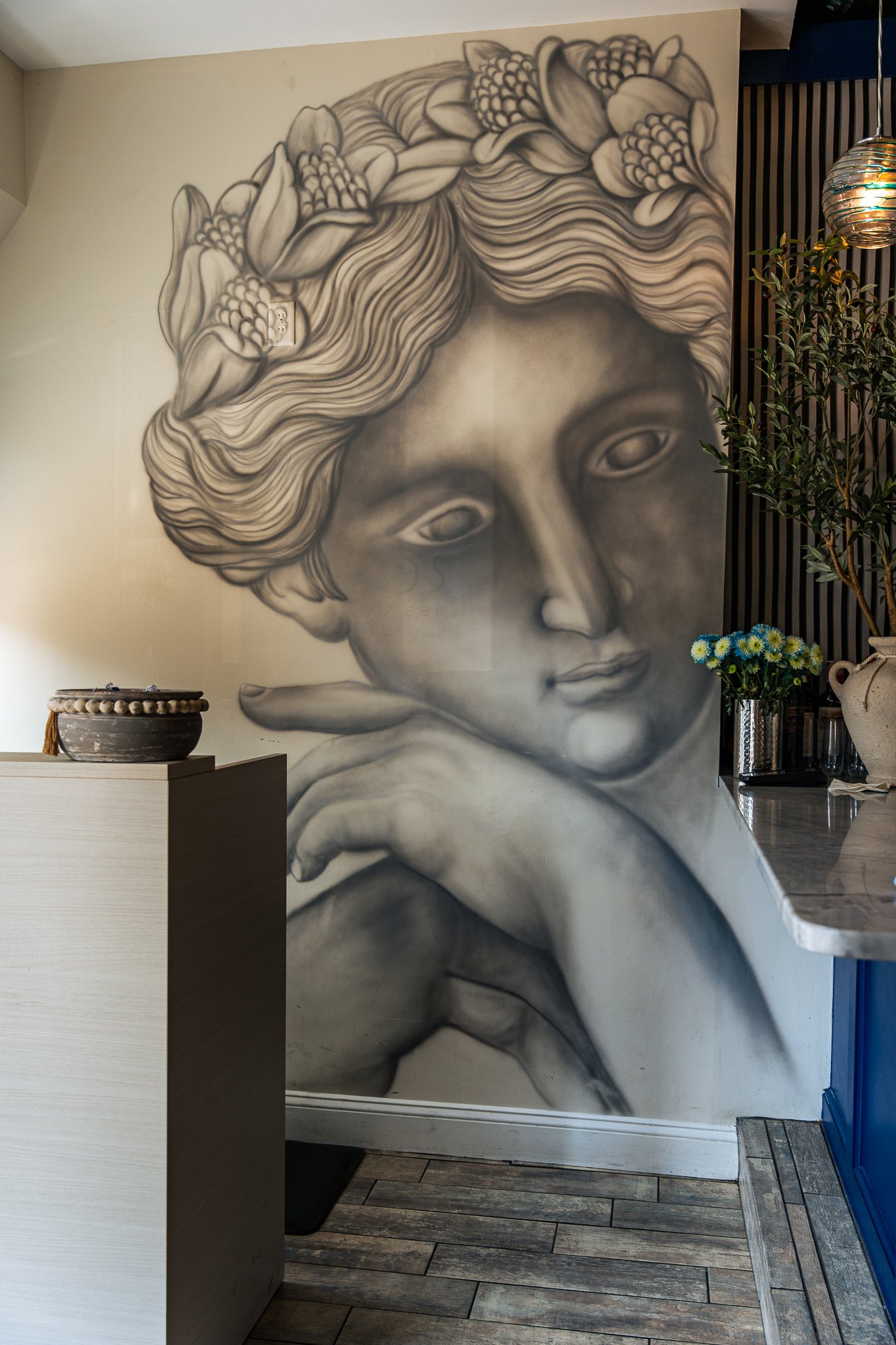 Black and white mural of a woman's face with her hand resting on her chin, surrounded by flowers in her hair, on a wall inside a room.