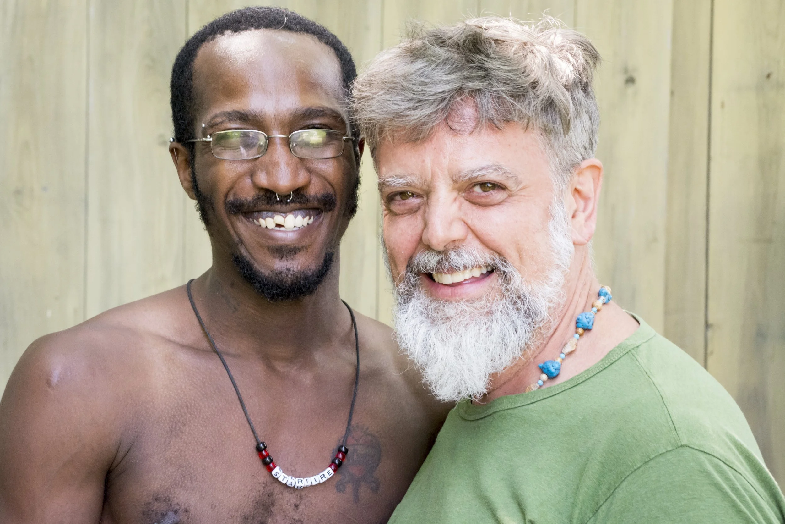 Two men smiling and embracing outdoors, one shirtless with glasses and a beaded necklace, the other wearing a green shirt and a beaded necklace, standing in front of a wooden fence.