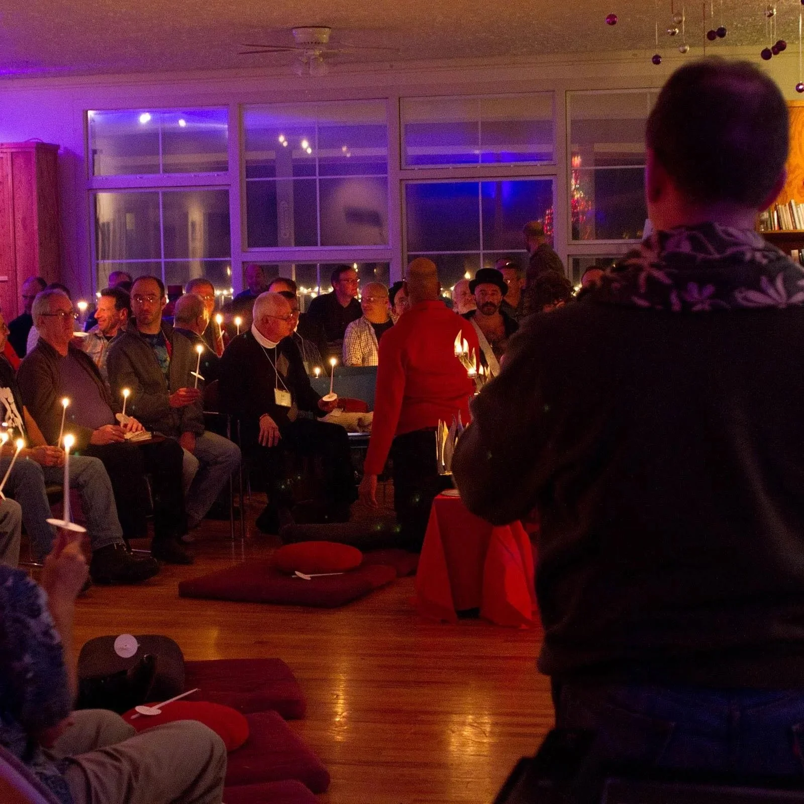 A group of people gathered in a living room during a candlelit event, with some seated on cushions and others standing, in front of large windows decorated for Christmas, with some wearing festive attire.