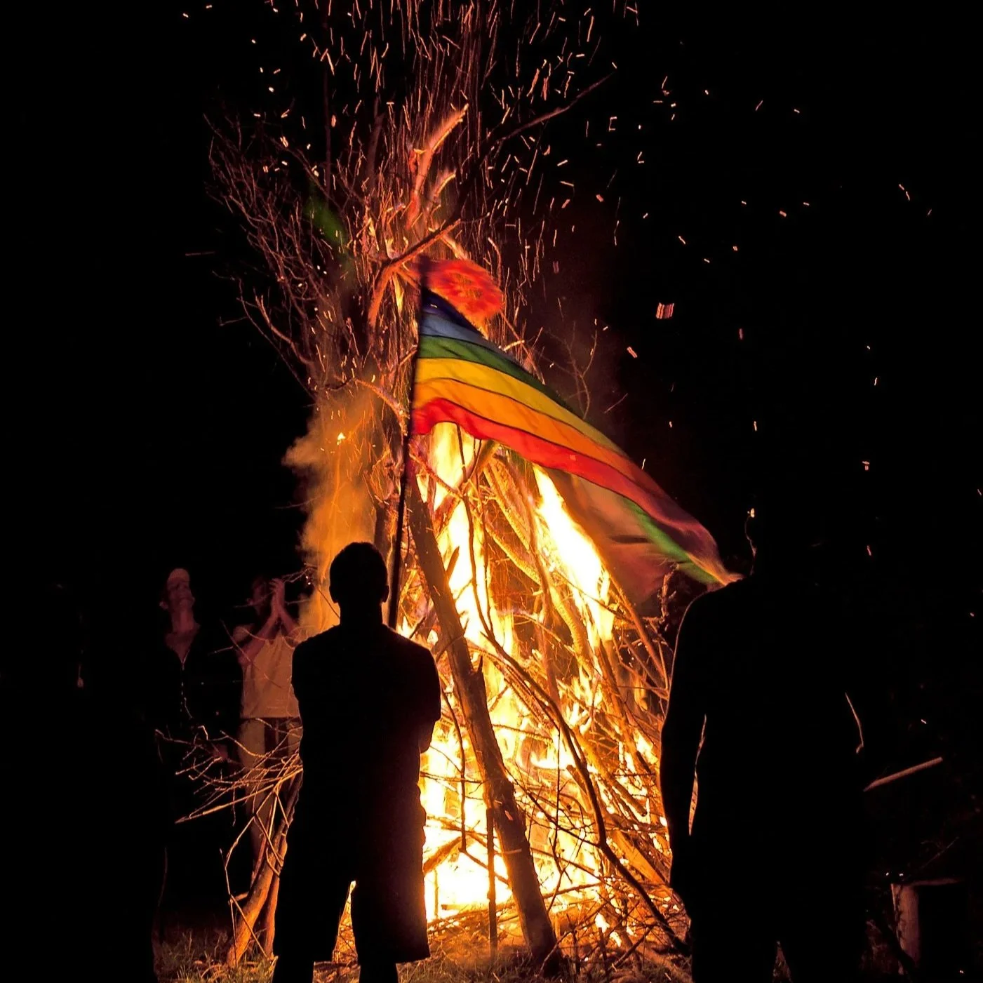 Silhouettes of three people watching a large bonfire with a multicolored flag and sparks flying in the night sky.