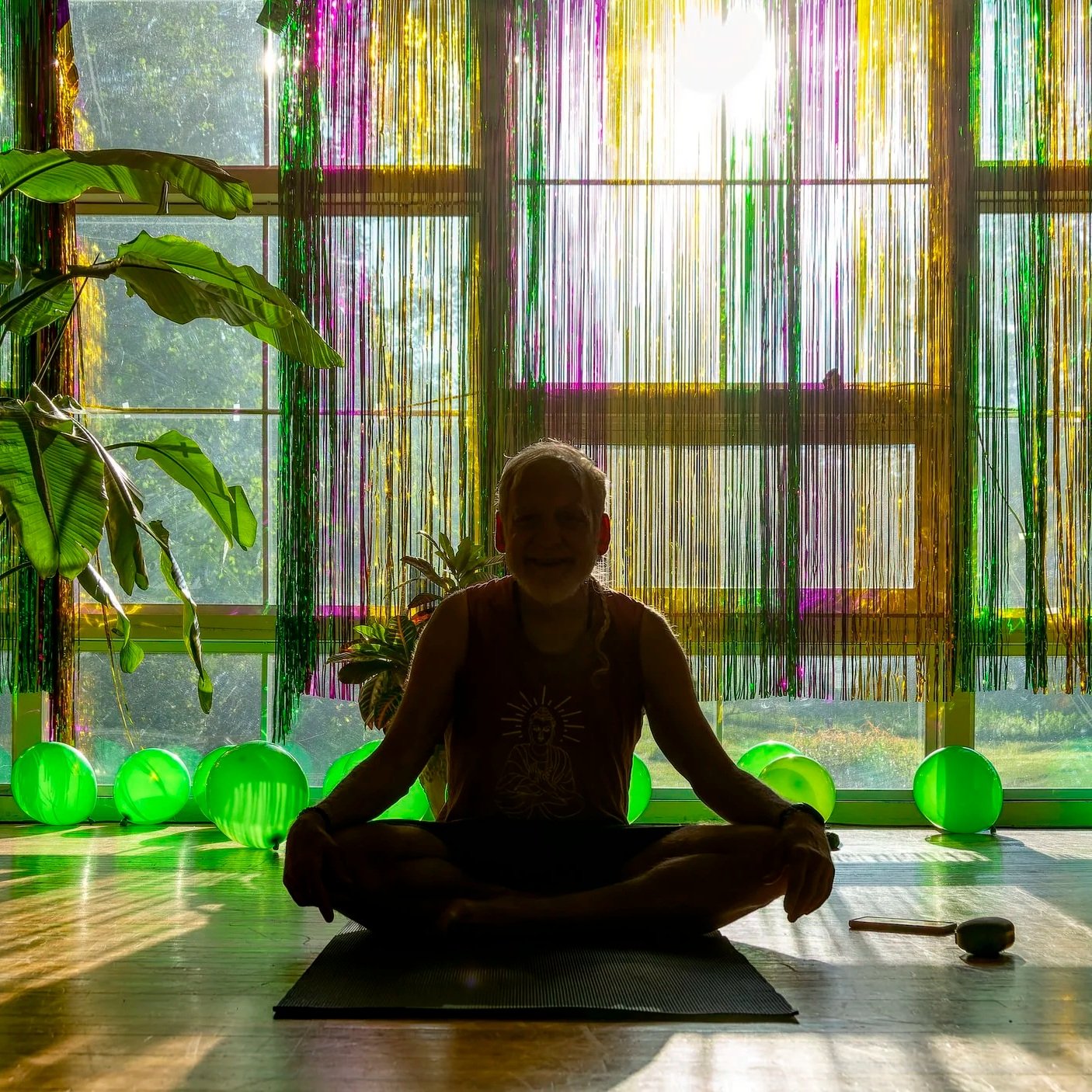 Person sitting cross-legged on yoga mat in a room with bright natural light coming through large windows, decorated with colorful streamers and green balloons, with houseplants nearby, in a yoga or meditation setting.