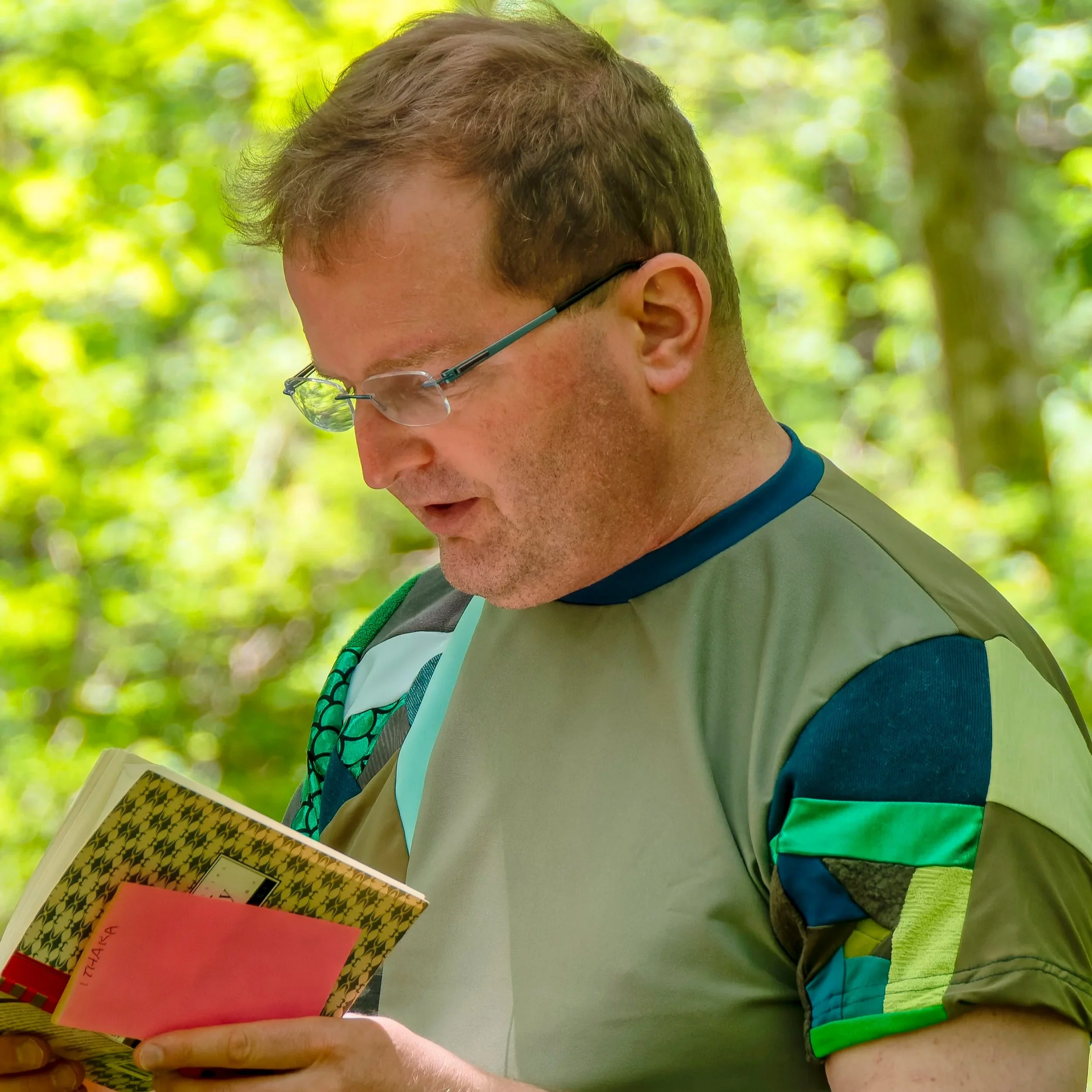 A person with short hair wearing glasses and a short-sleeved shirt, sitting outdoors with a blurred green background, holding a colorful book or magazine.