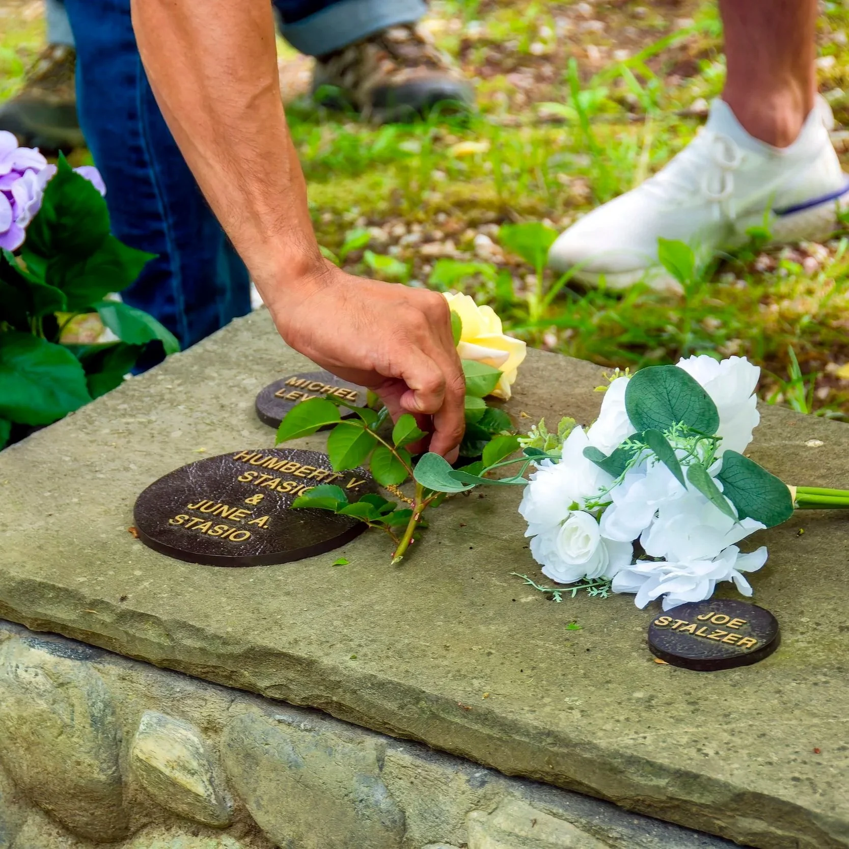 Person placing flowers at a memorial with engraved names, surrounded by other flowers and greenery.