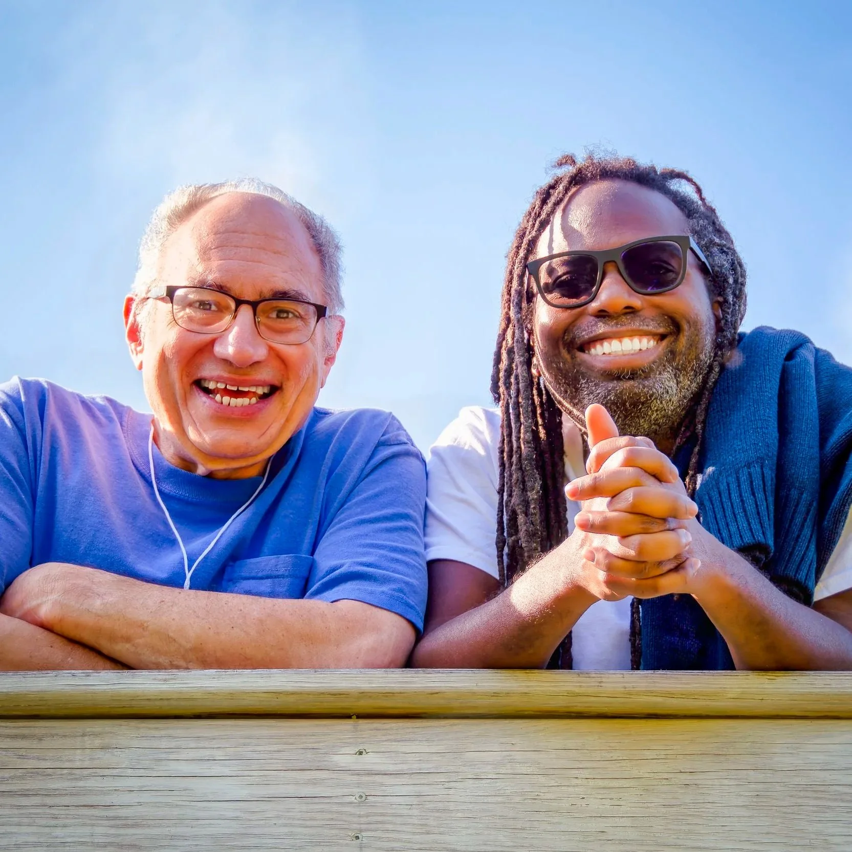 Two smiling men, one with gray hair and glasses, the other with dreadlocks and sunglasses, taking a selfie outdoors against a clear blue sky.