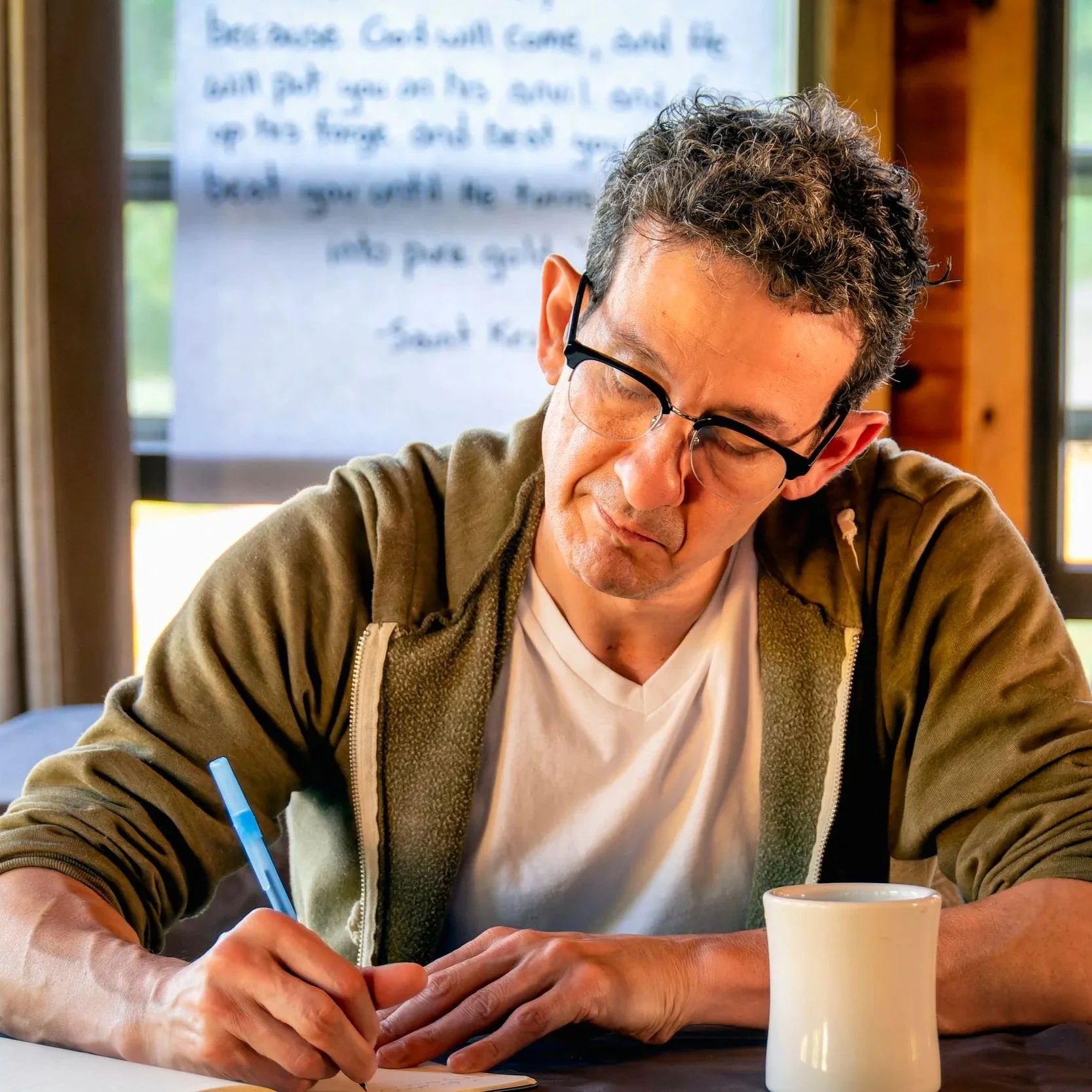 A man with curly hair and glasses, wearing a beige zip-up jacket, is sitting at a table writing with a blue pen in a notebook. There is a white mug in front of him, and he is in a room with wood-paneled walls and windows. A blurred piece of paper wit