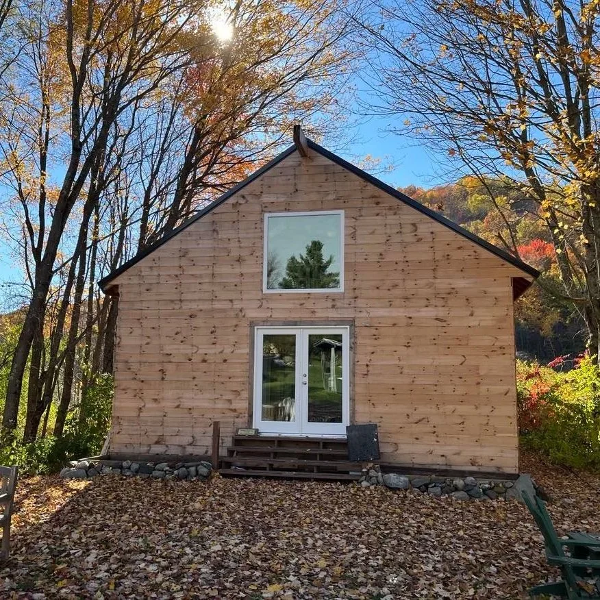 A wooden house with a gabled roof and a glass door, surrounded by autumn trees with colorful leaves, on a clear day.