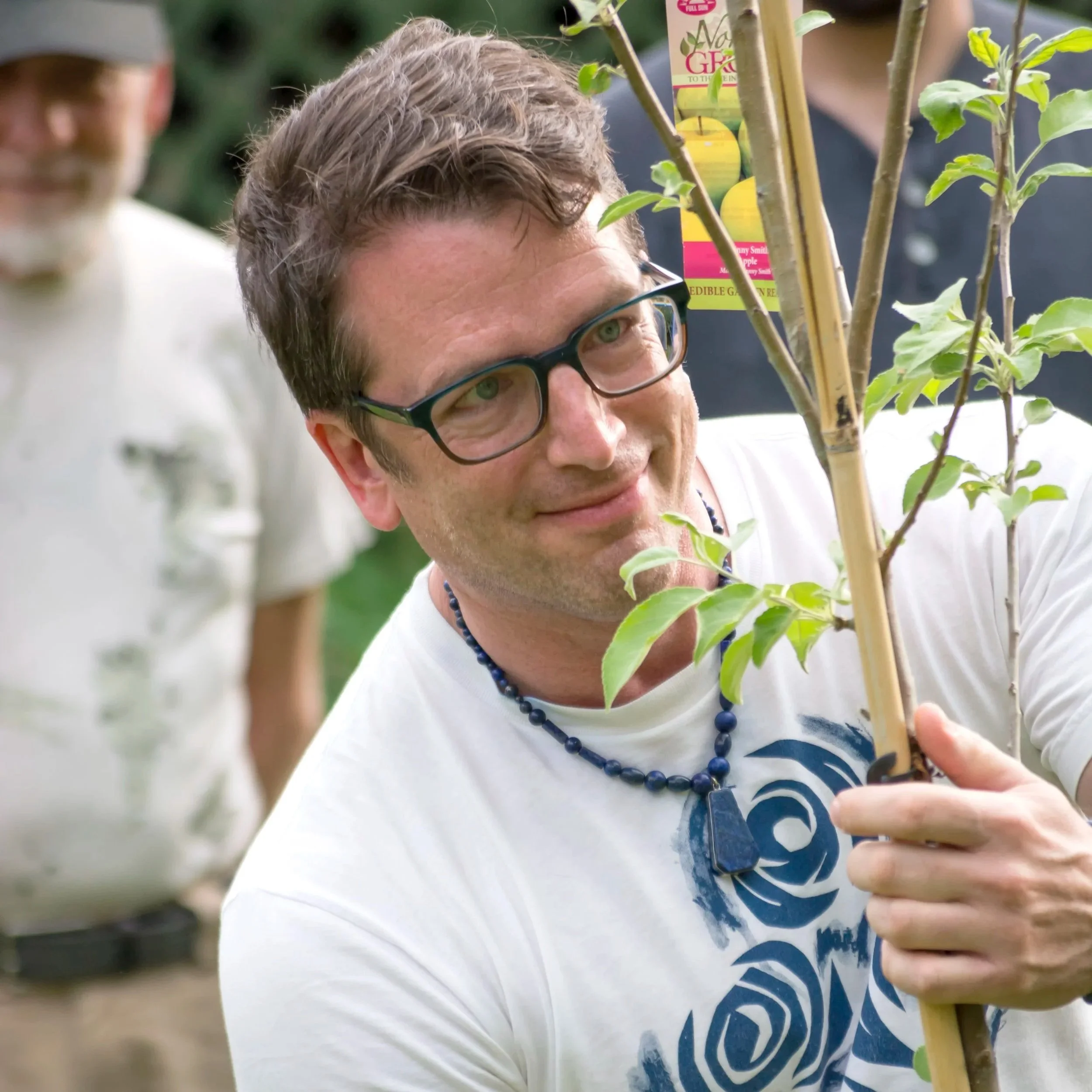 A man with glasses and a beaded necklace inspecting a young tree outdoors, with another person blurred in the background.