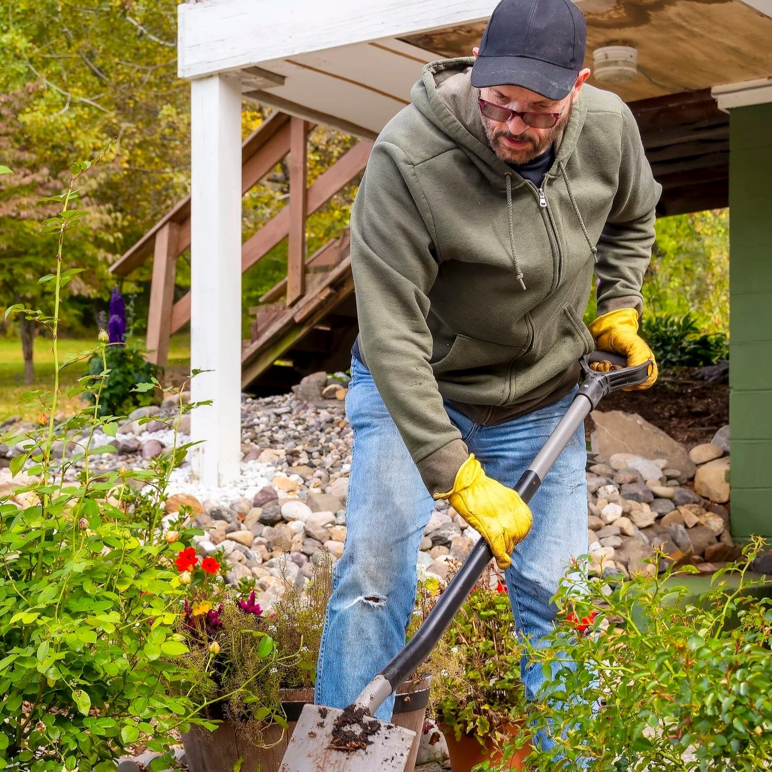 A man in a black cap, hoodie, and yellow gloves is gardening with a shovel amidst colorful flowers in a backyard.