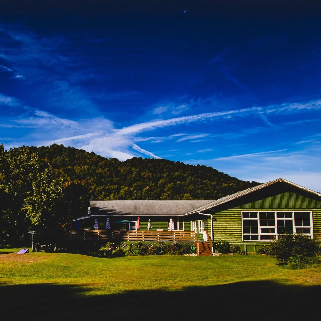 Green house with a spacious deck, umbrellas, and chairs, set against lush green hills and a vibrant blue sky with wispy clouds and a visible moon.