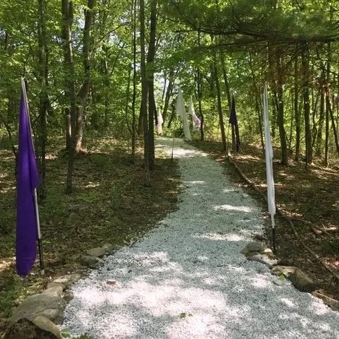 A gravel pathway through a wooded area lined with purple and white flags.