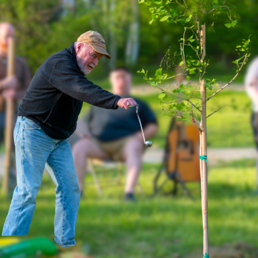An elderly man wearing a tan cap, black jacket, and jeans is playing golf with a golf club on a grassy area. There are trees in the background and some people sitting on chairs, watching.