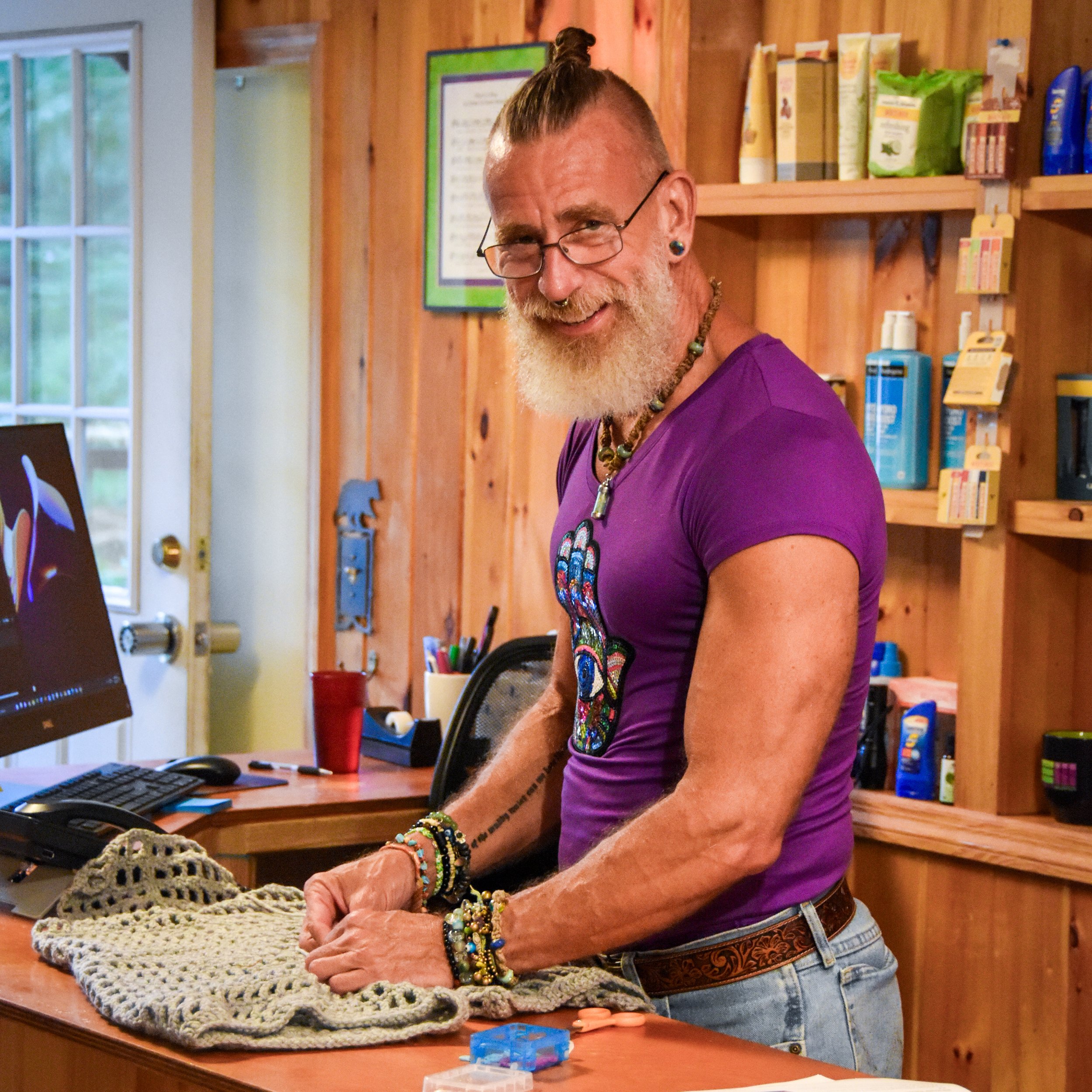 A smiling man with glasses, earrings, and colorful jewelry is standing at a table, working on a crochet project. He is wearing a purple shirt and denim jeans with a patterned belt. The setting is a cozy wooden room with bookshelves and a computer.