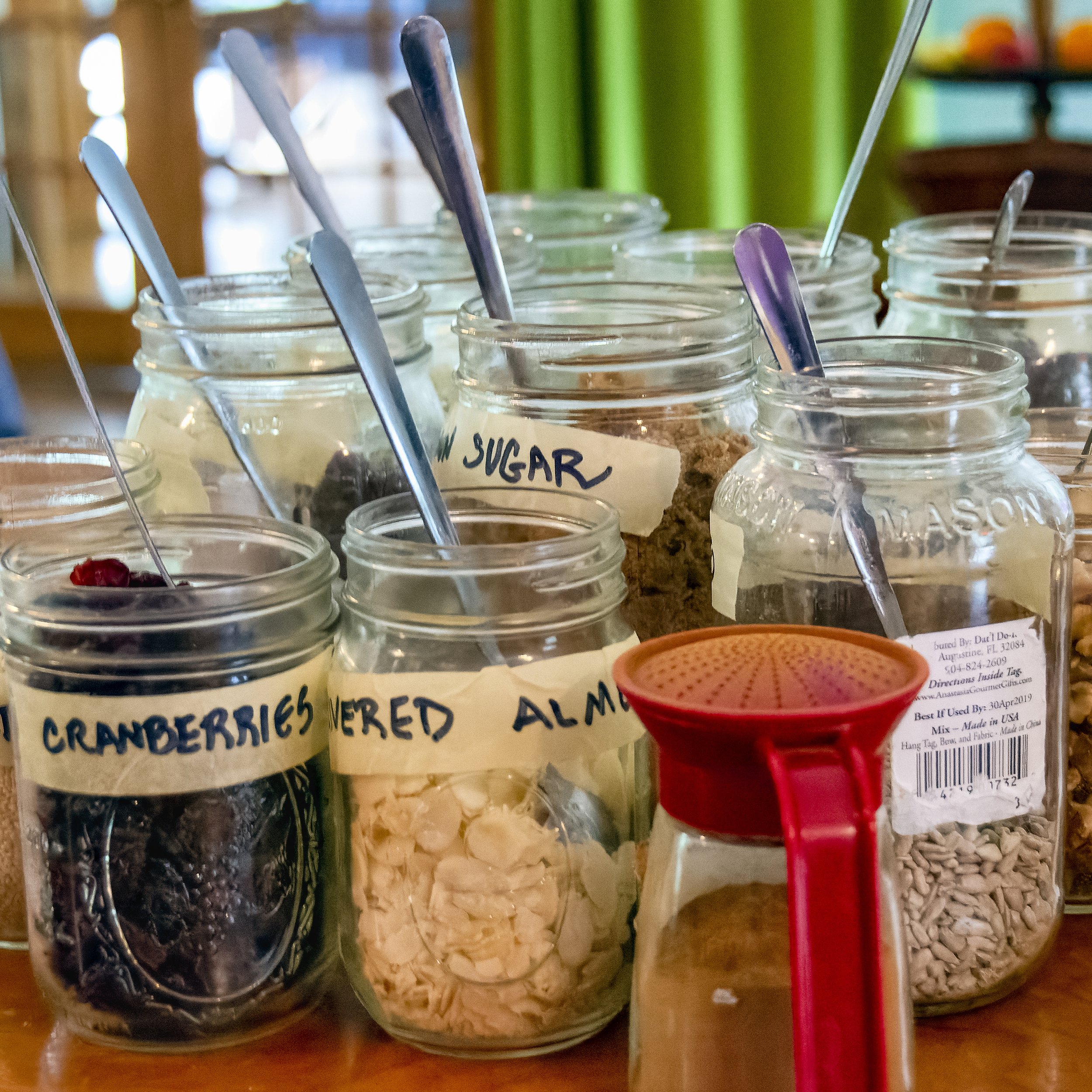 Assorted jars of flavored powders and sprinkles, including cranberry, almond, and vanilla, with a red spoon.