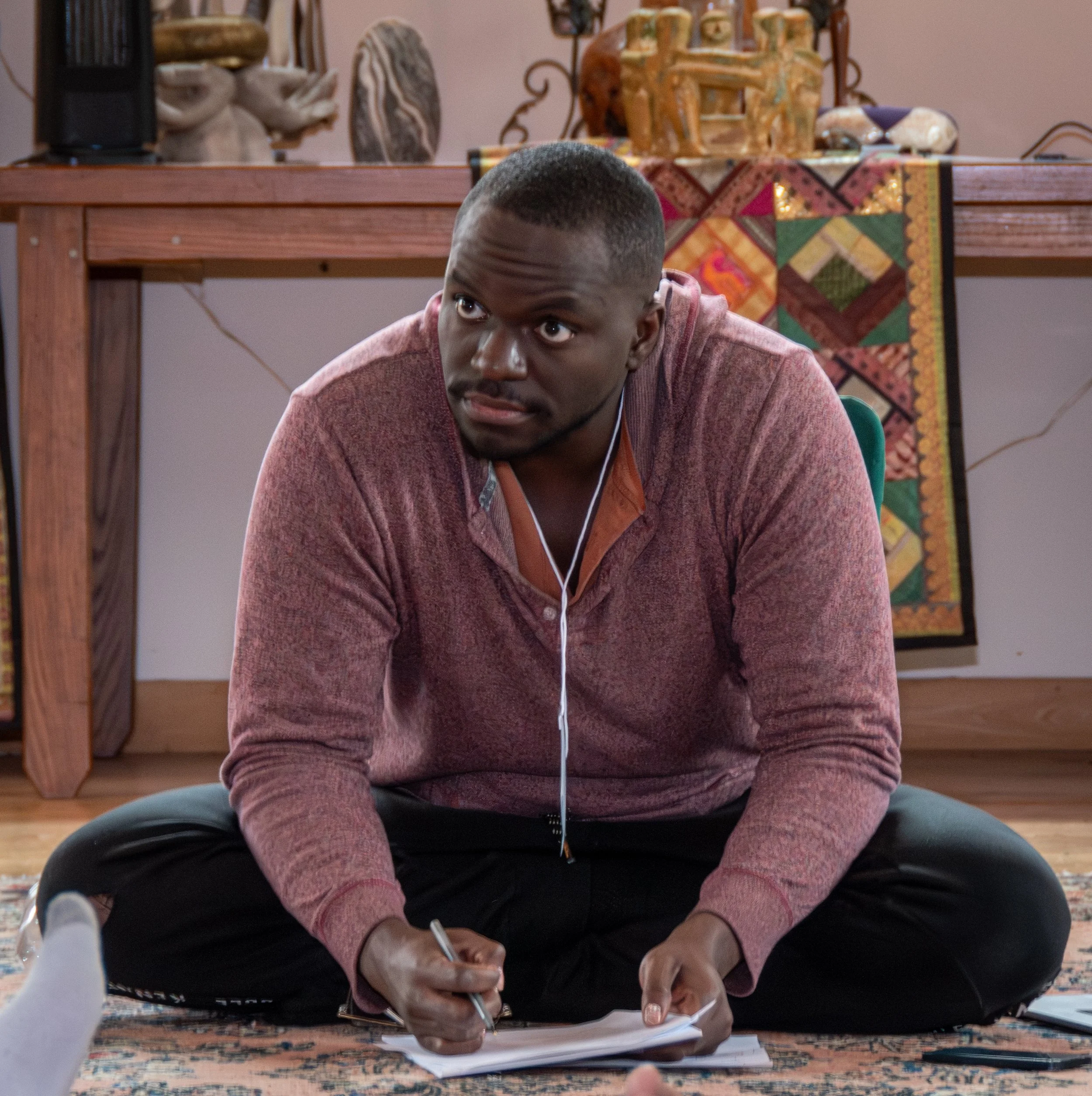 A young man with short hair, wearing a pinkish-red jacket, is sitting on a patterned rug on the floor, writing on a notepad with a pen. He appears to be focused, and he has white earphones in his ears. Behind him, there is a wooden table with decorat