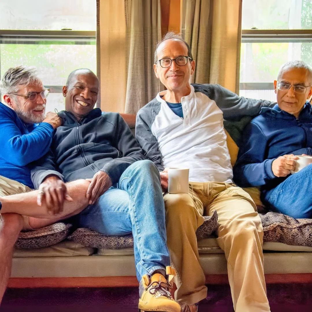 Four men sitting on a couch in a room with large windows and curtains, smiling and holding mugs.