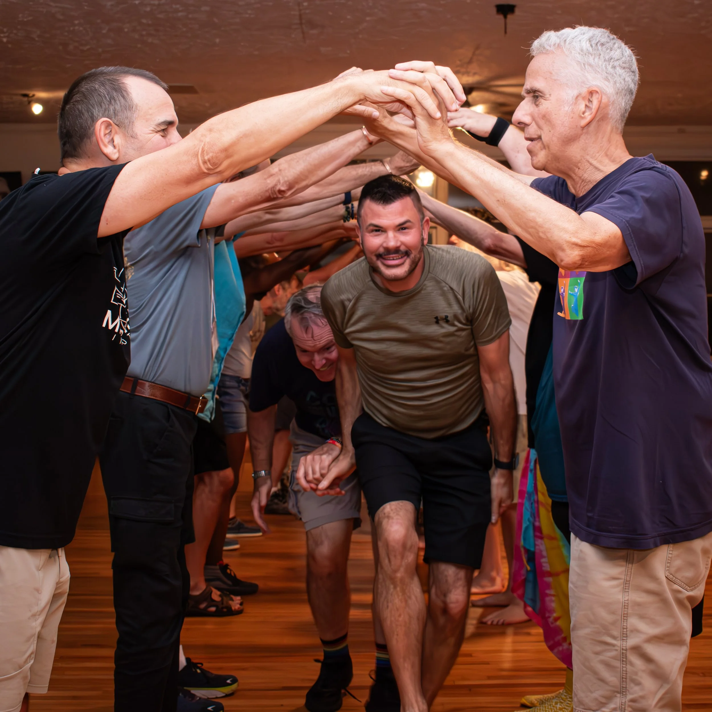 A group of people participating in a team-building activity, forming an arch with their hands and supporting a man with a beard and shorts as he crawls under the arch.