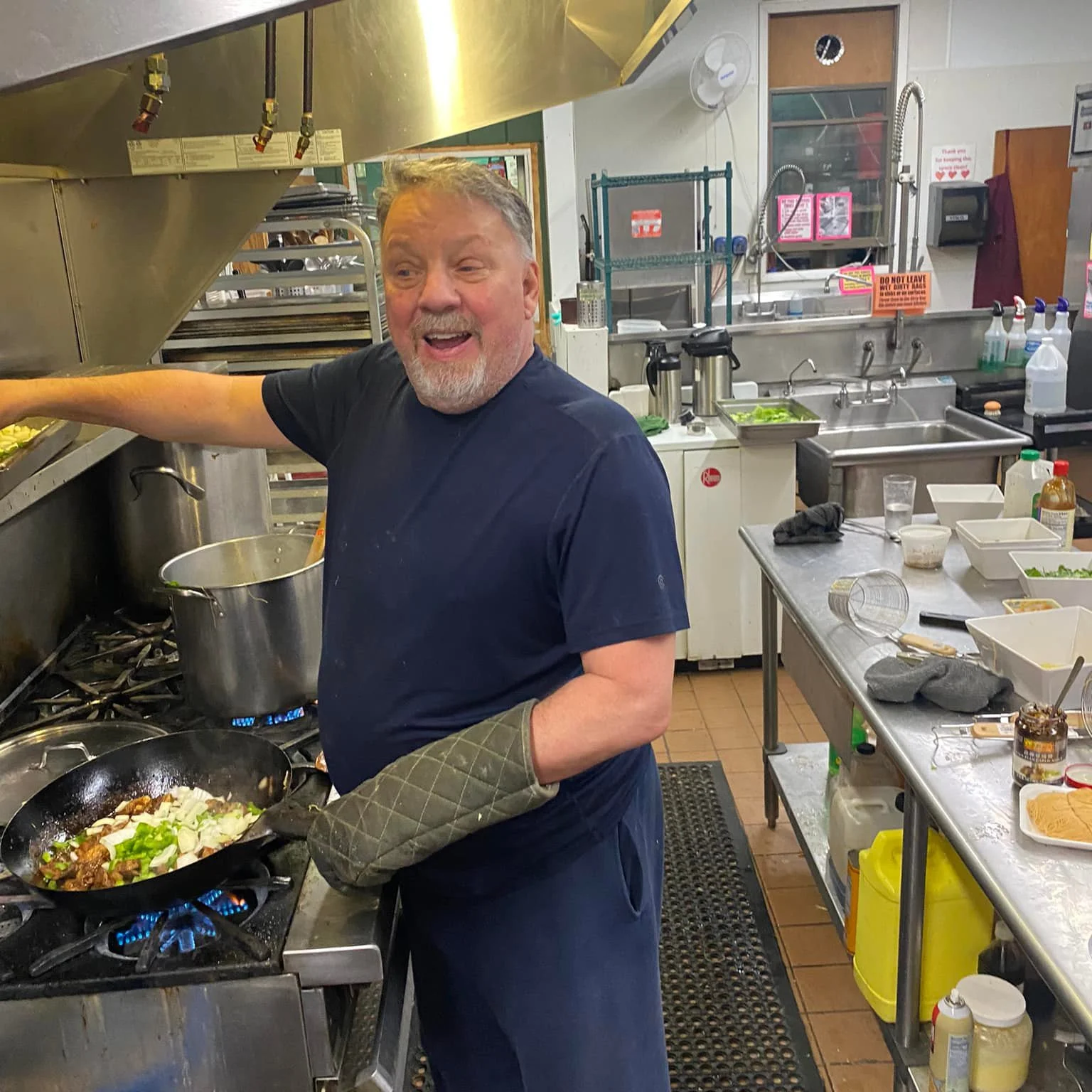 A man is cooking in a commercial kitchen, stir-frying vegetables and meat in a black wok on a stove. He is smiling and looking at the camera. The kitchen has stainless steel countertops and shelves with various cooking supplies and ingredients visibl