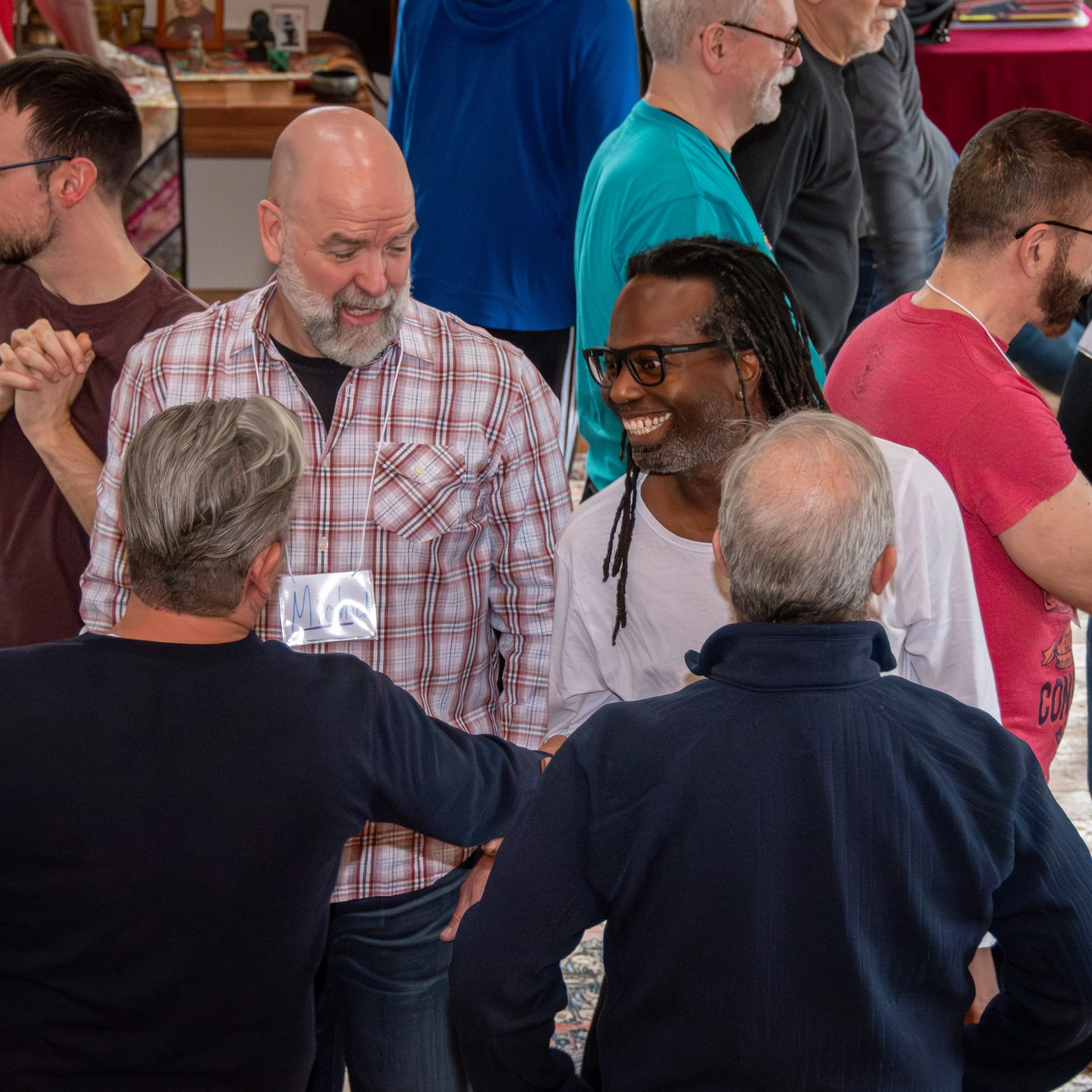 A group of diverse people standing closely together at a gathering, seen from the side, some smiling and talking, in an indoor setting with a patterned carpet and furniture in the background.