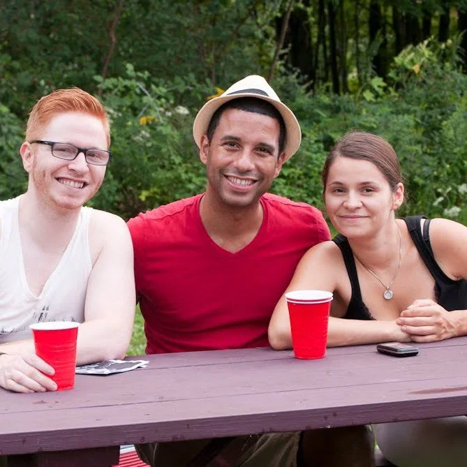 Three friends sitting at an outdoor picnic table, smiling, with greenery in the background. One holds a red cup, and there are additional red cups on the table.