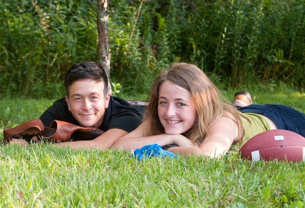 Two teenagers lying on the grass in a park with trees in the background, smiling at the camera. A football is nearby.