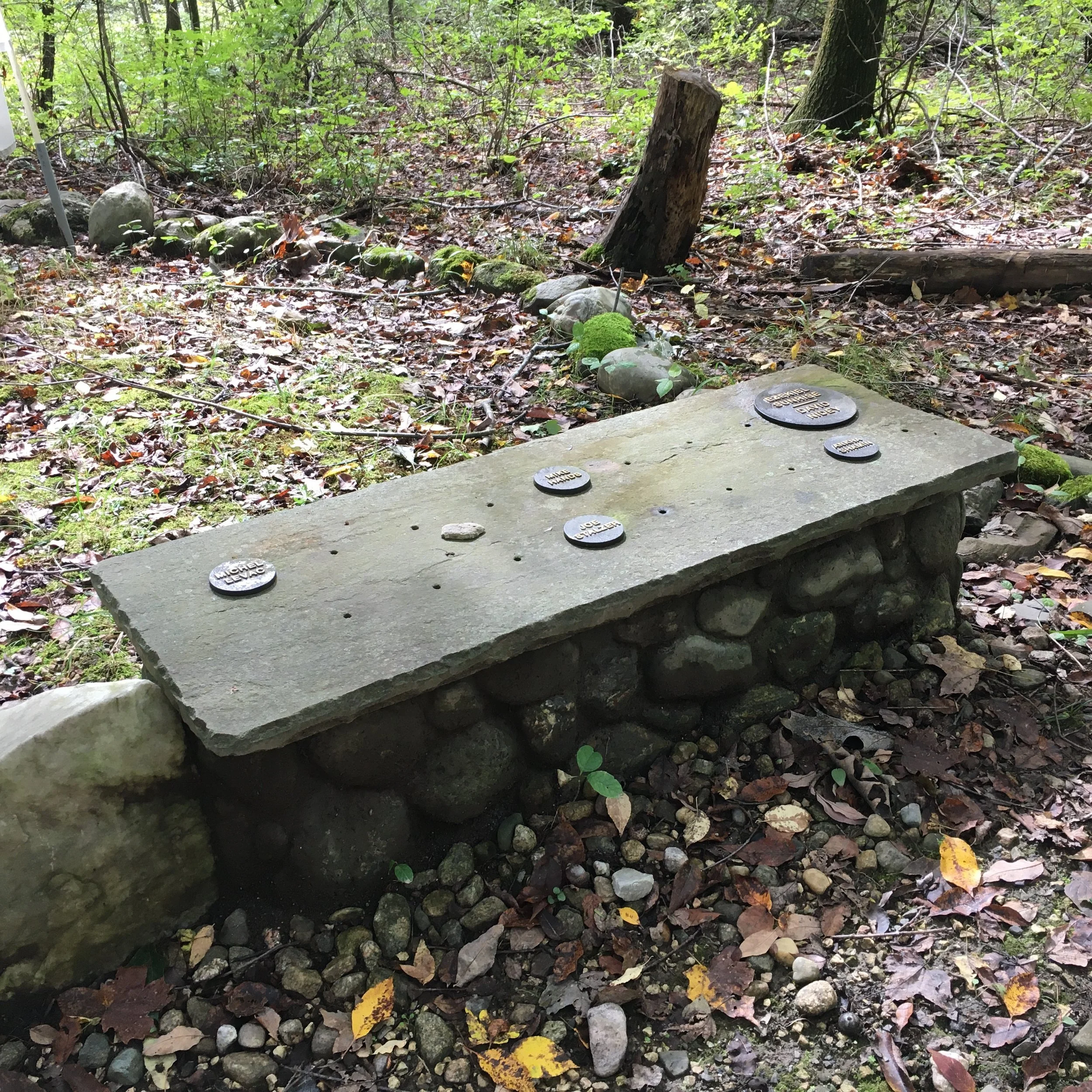 A small stone and metal memorial bench in a wooded area surrounded by leaves and rocks, with engraved metal plaques on top.