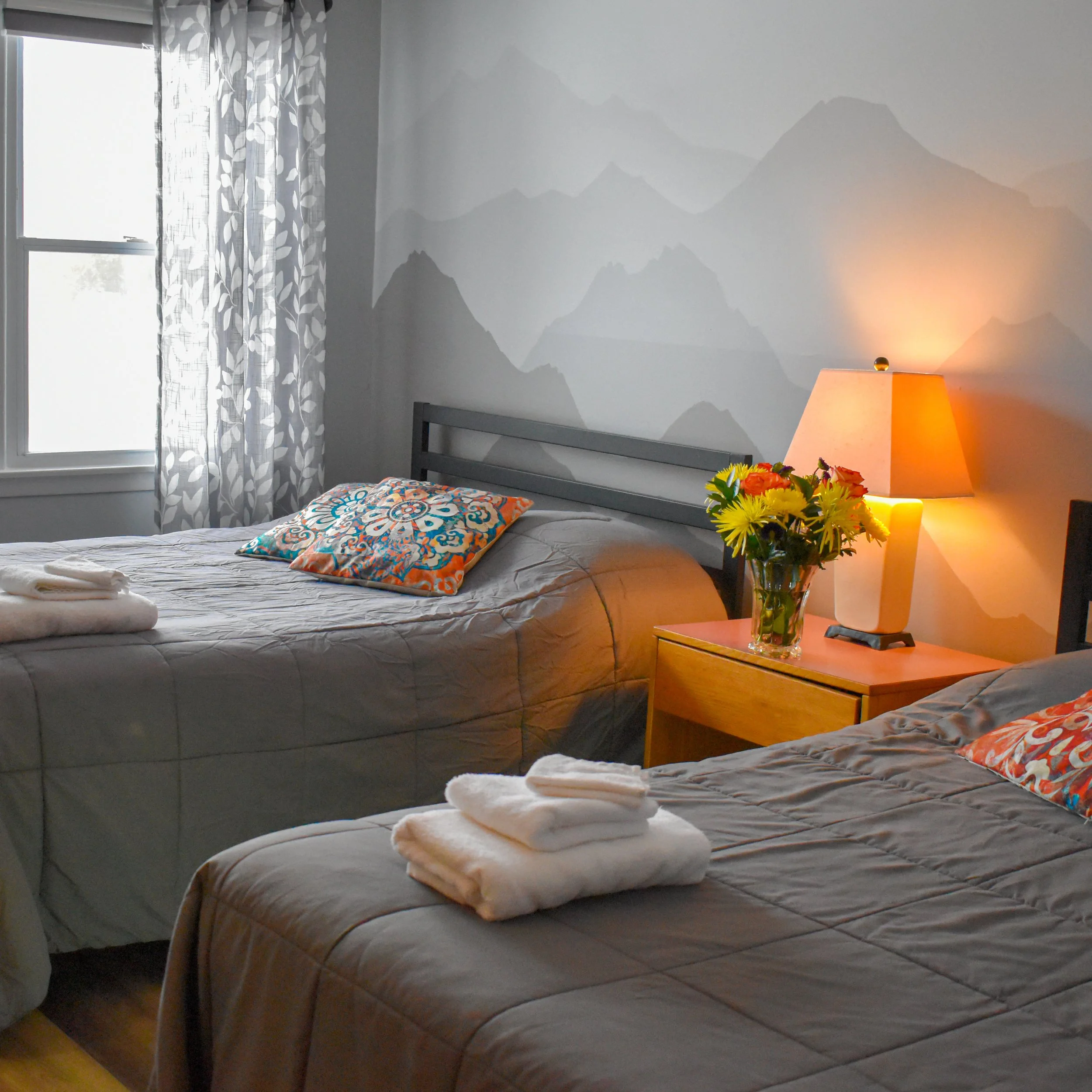 A bedroom with a window, a bed with a colorful pillow, a wooden nightstand with a lamp and a vase of yellow flowers, and mountain-themed wall art.