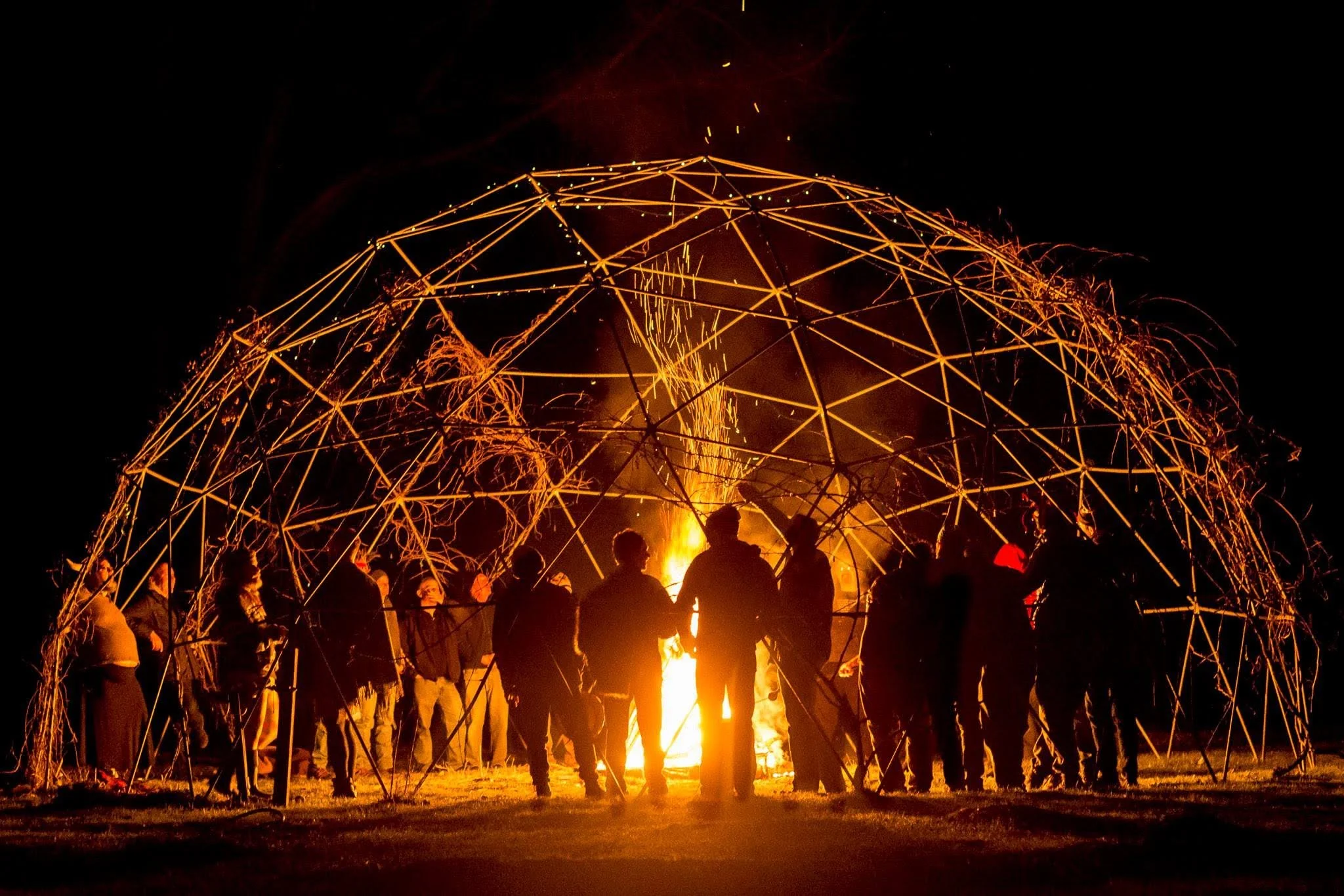 People gathered around a large bonfire beneath a geometric metal sculpture structure at night.
