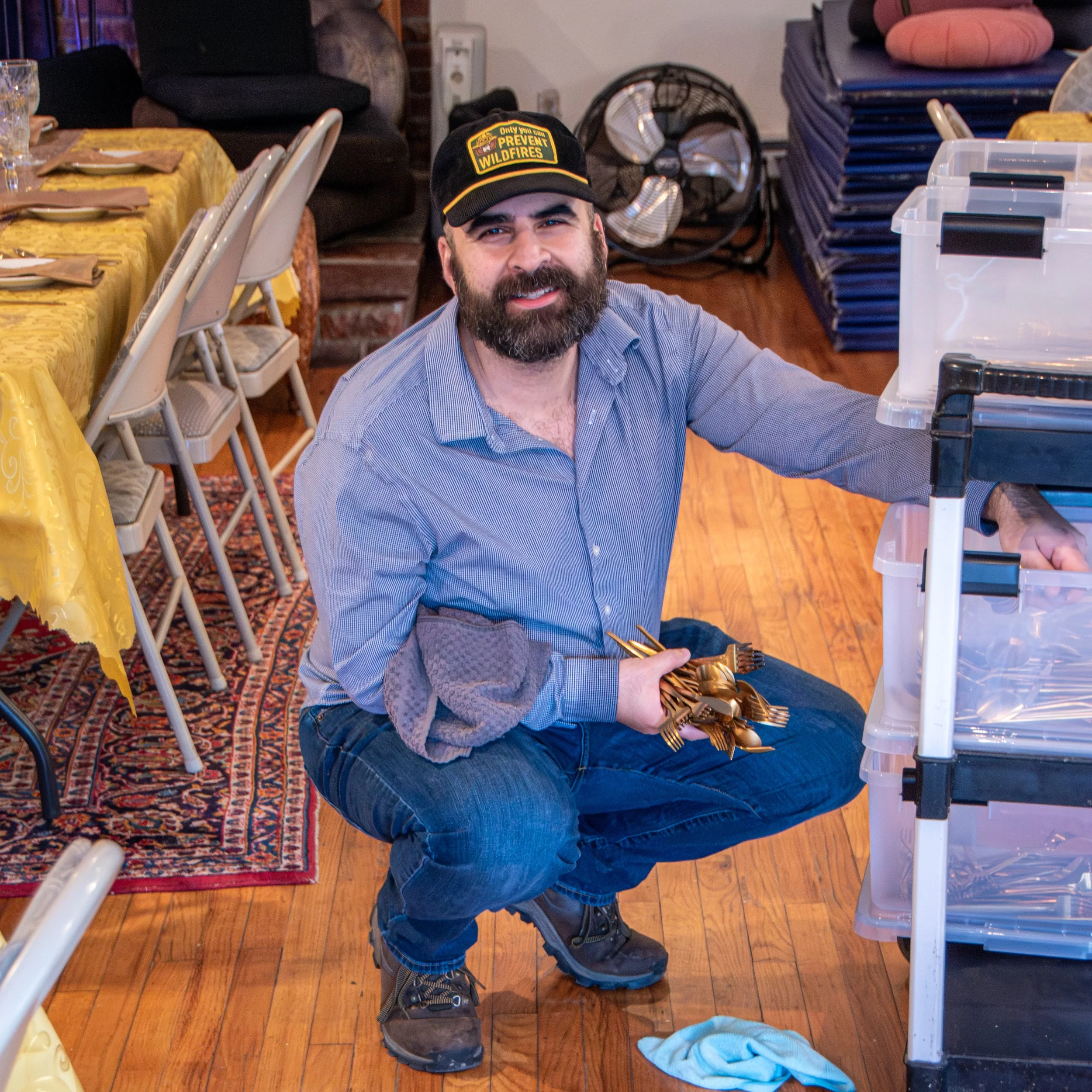 A man with a beard, wearing a black cap and blue shirt, is sitting on the floor, smiling, inside a room with tables, chairs, and storage containers.