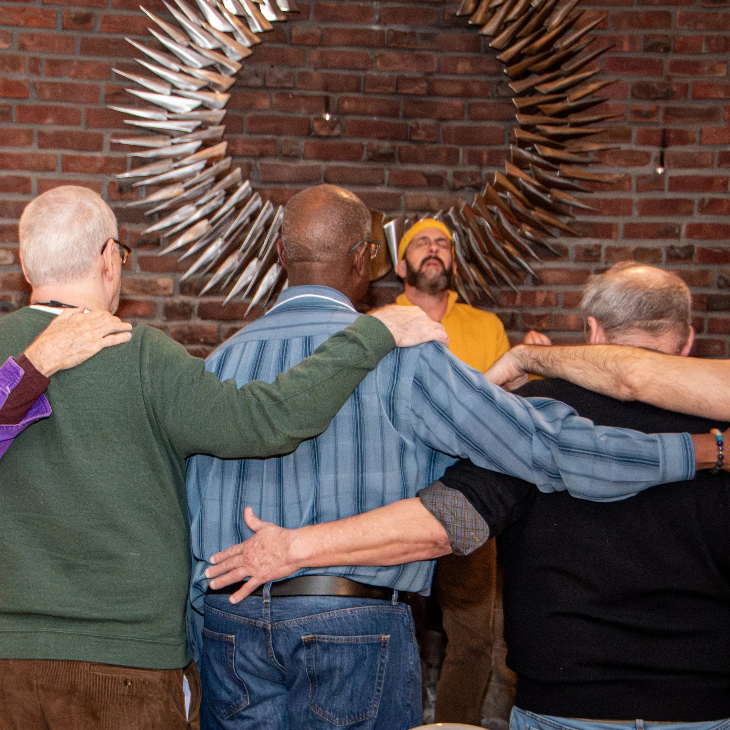 People standing with arms around each other's shoulders in front of a brick wall, participating in a group prayer or moment of unity.