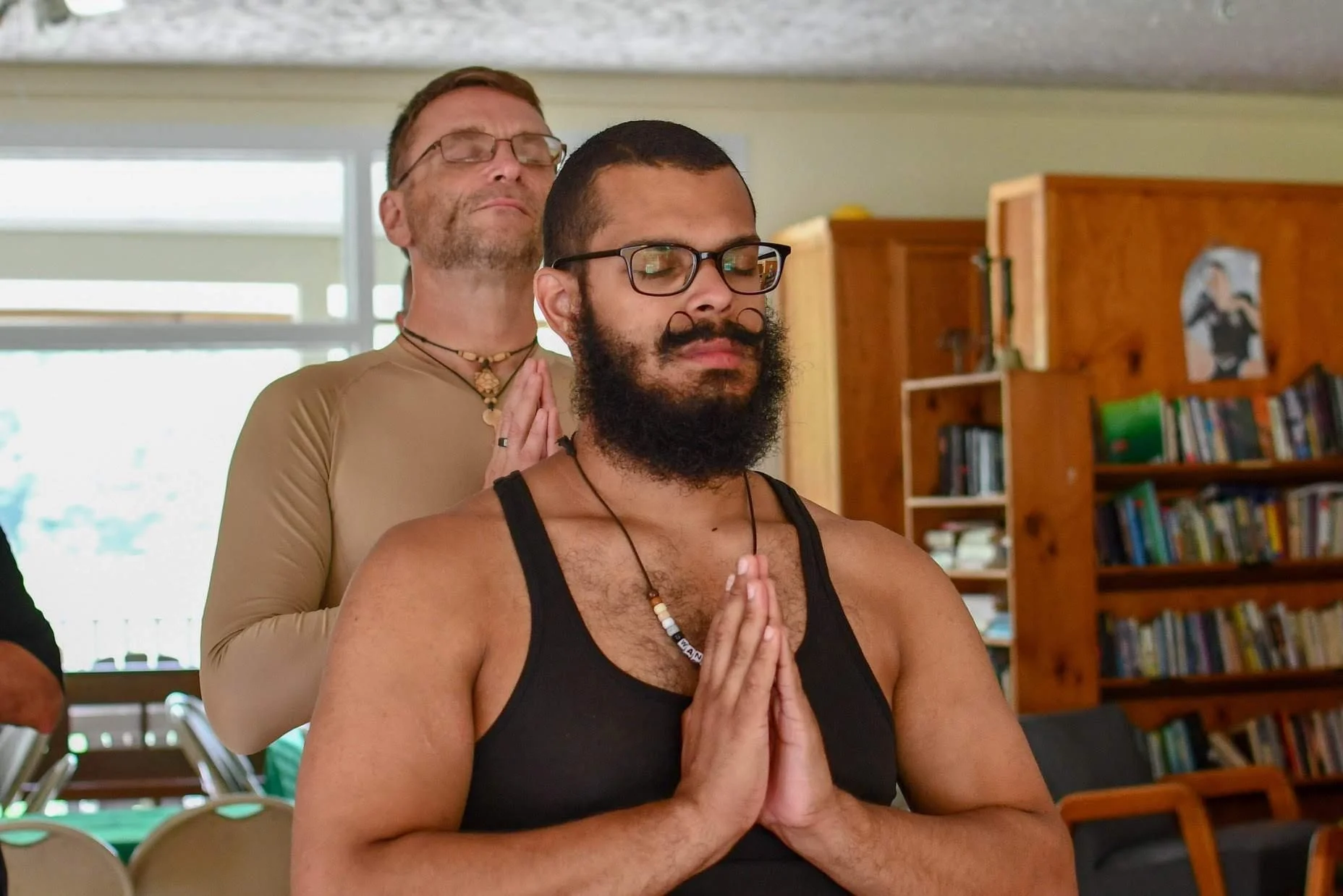 Two men meditating indoors with closed eyes and hands in prayer position, one standing behind the other.
