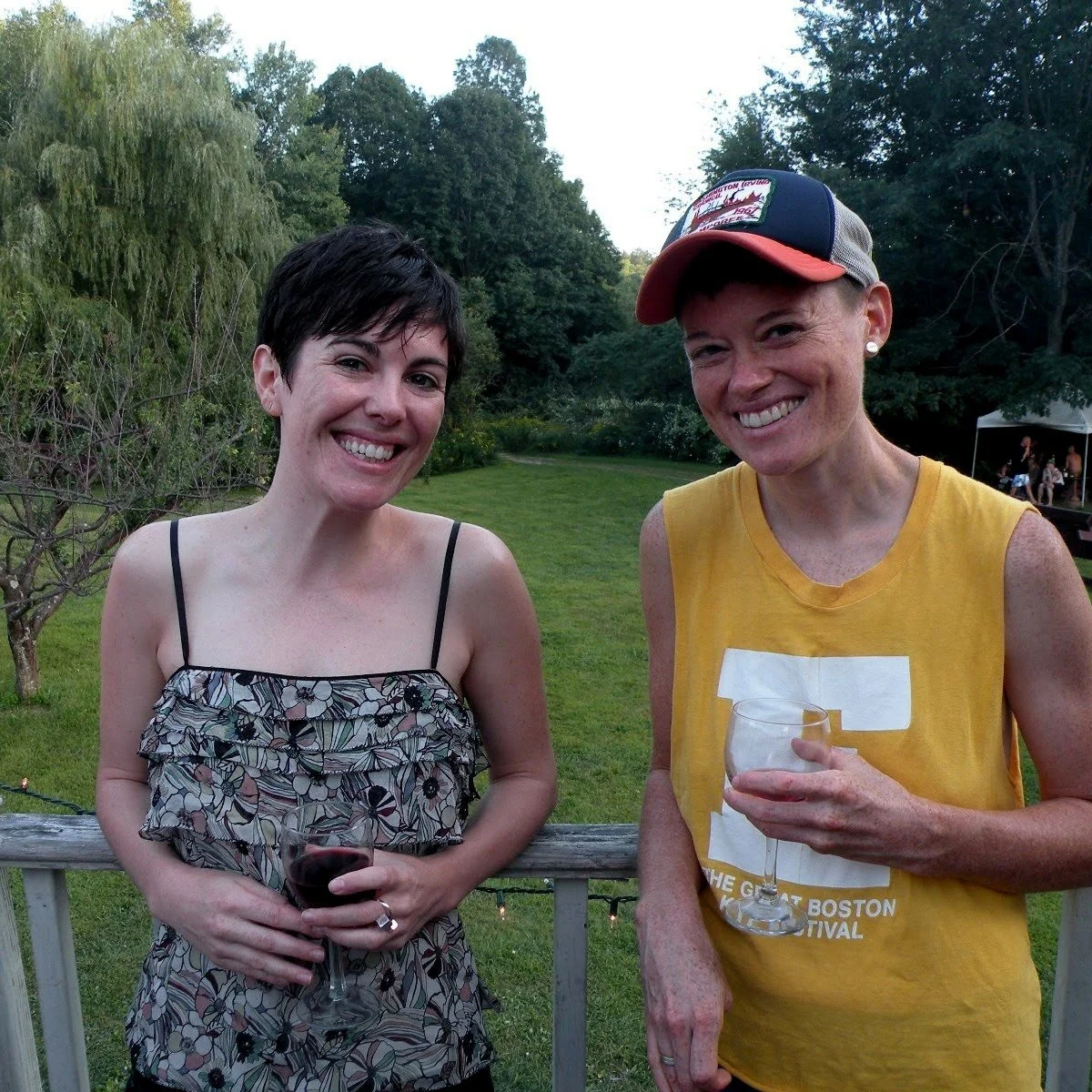 Two women smiling and holding glasses of wine outdoors on a wooden deck, with green trees and a lawn in the background.