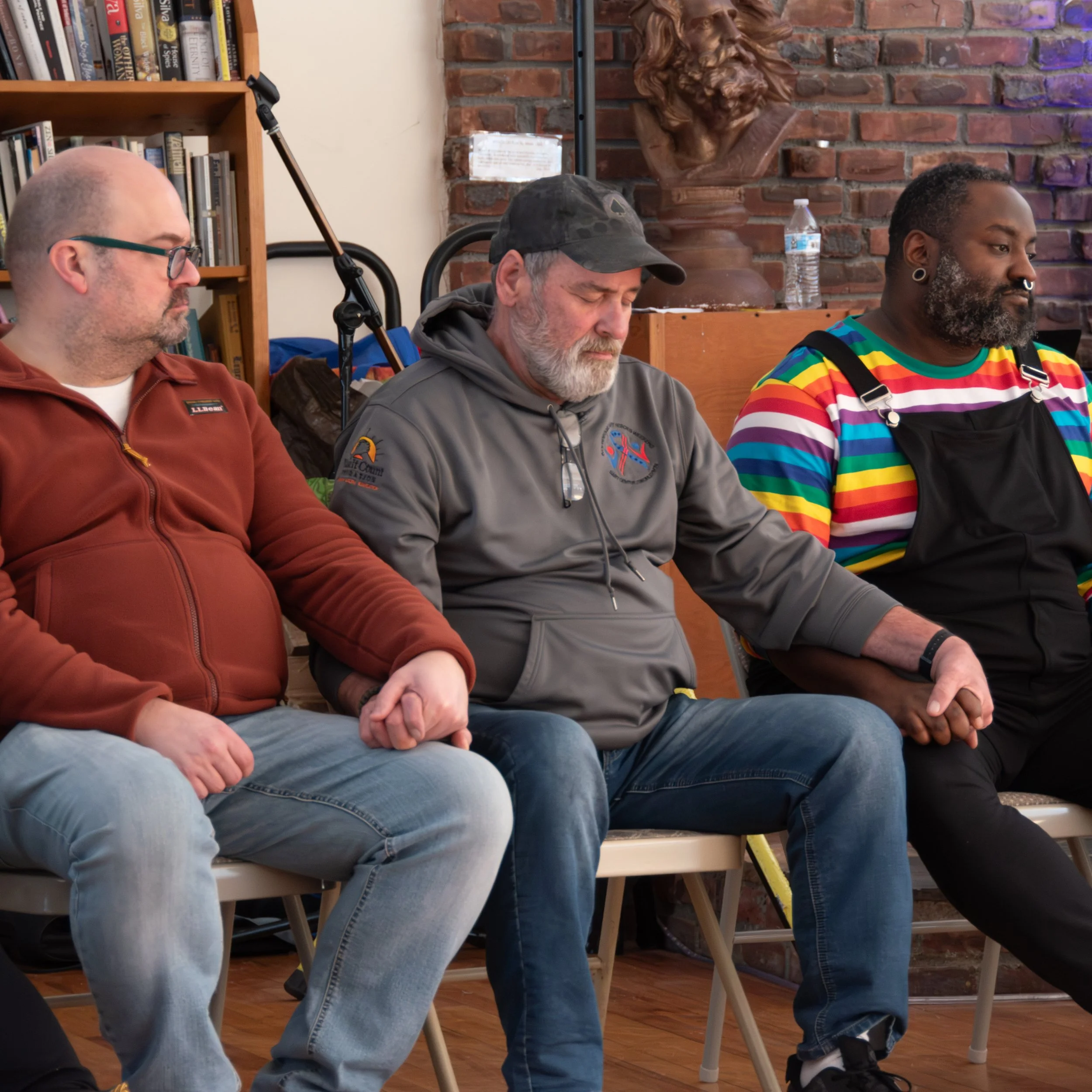 Three men sitting on chairs with their hands folded, eyes closed, possibly participating in a meditation or prayer session, in a room with a brick wall, a bookshelf, and a wooden sculpture of a lion's head in the background.
