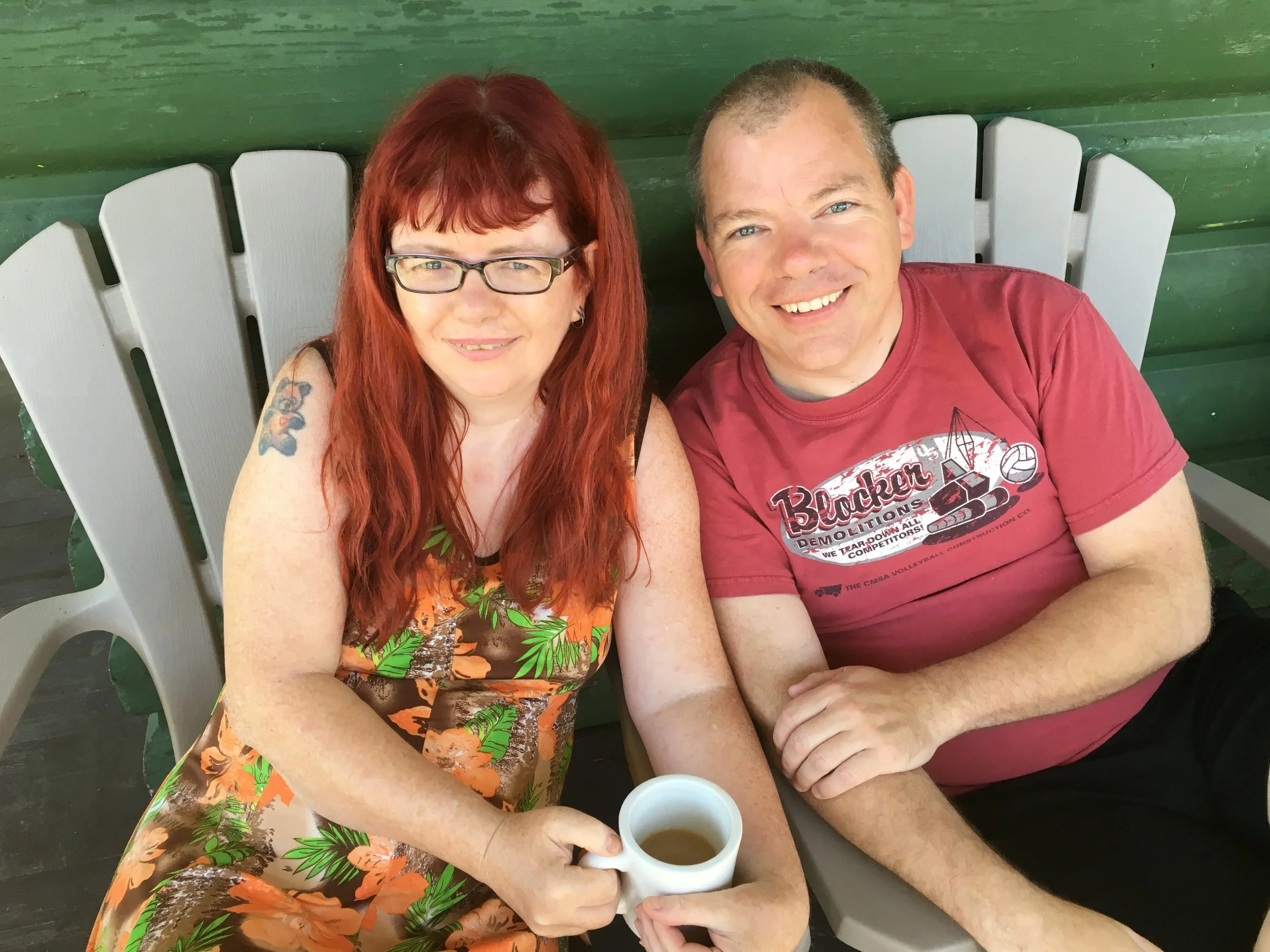 A woman with red hair and glasses holding a white mug, sitting beside a man with light hair wearing a red t-shirt, sitting on a porch with green wooden walls.