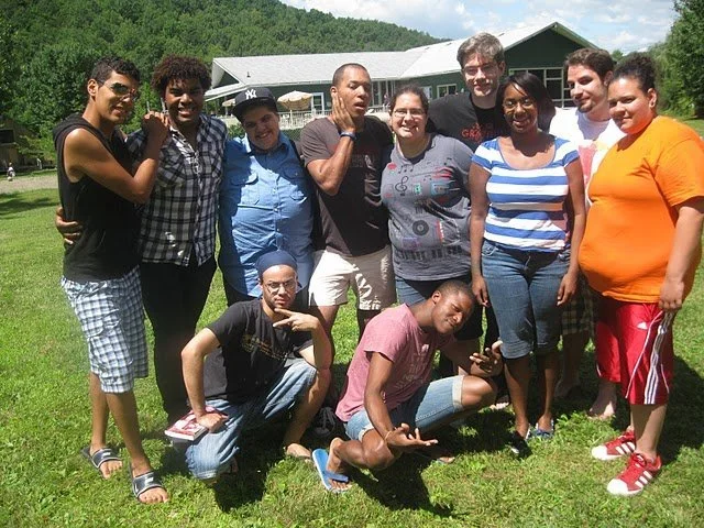 Group of ten young people standing and sitting on grass outdoors with a building and trees in the background.