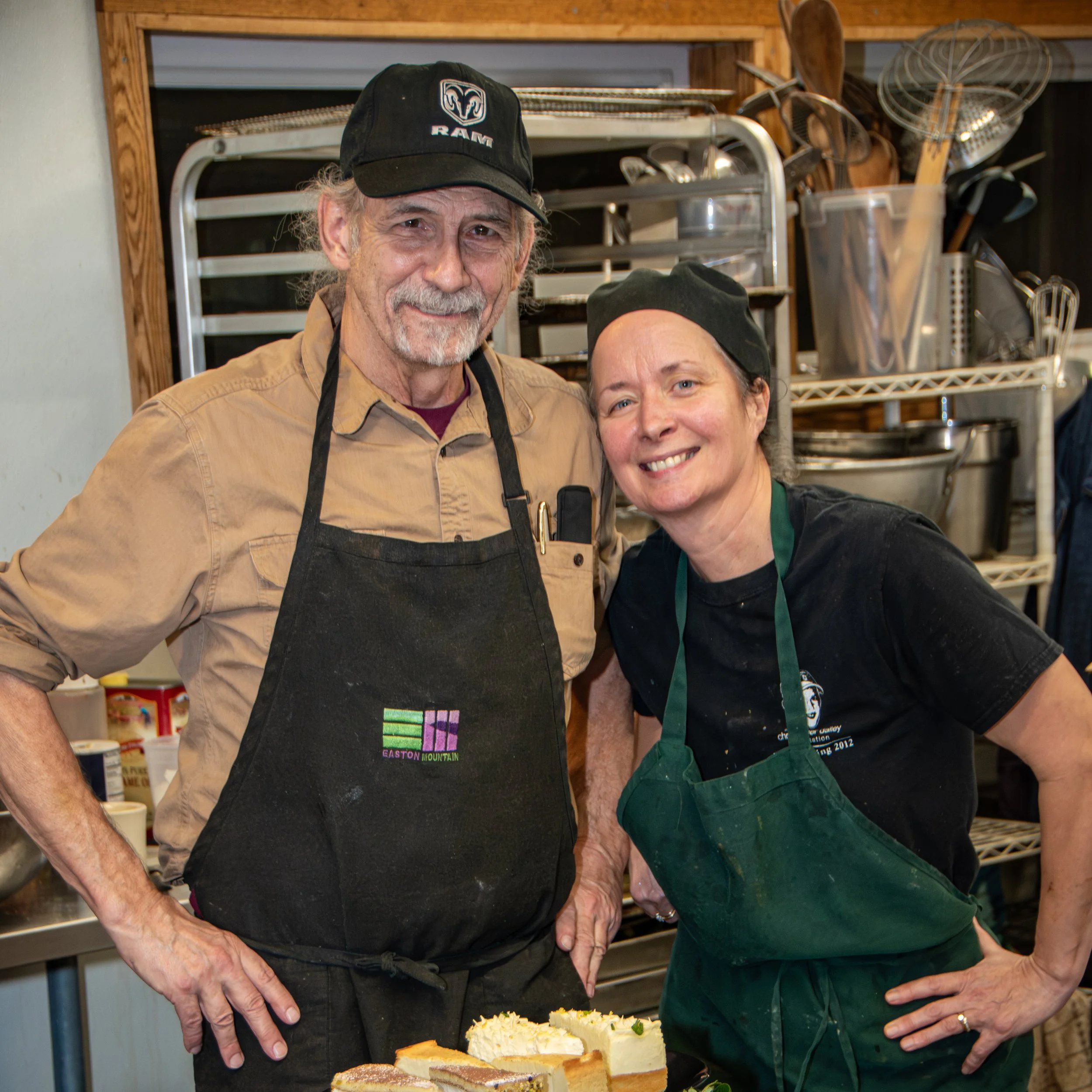 Two smiling people, a man and woman, in a kitchen with bake goods on a table in front of them, standing close together, wearing aprons and hats.