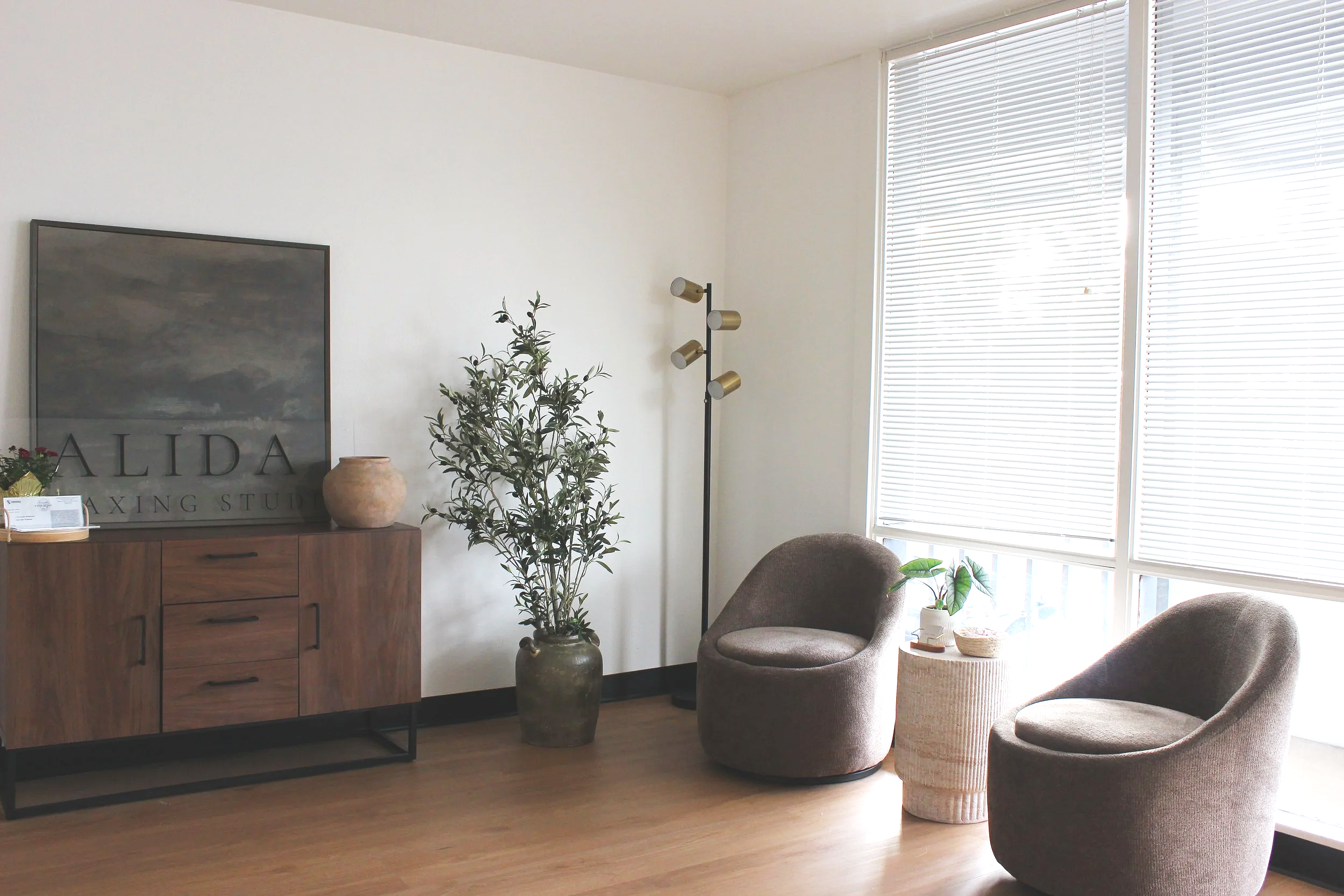 Interior of a waxing studio with a massage table, white armchair, side table with hand sanitizer, floor lamp, abstract black wall art, and a sign that reads 'ALIDA A WAXING STUDIO' on white paneled wall.