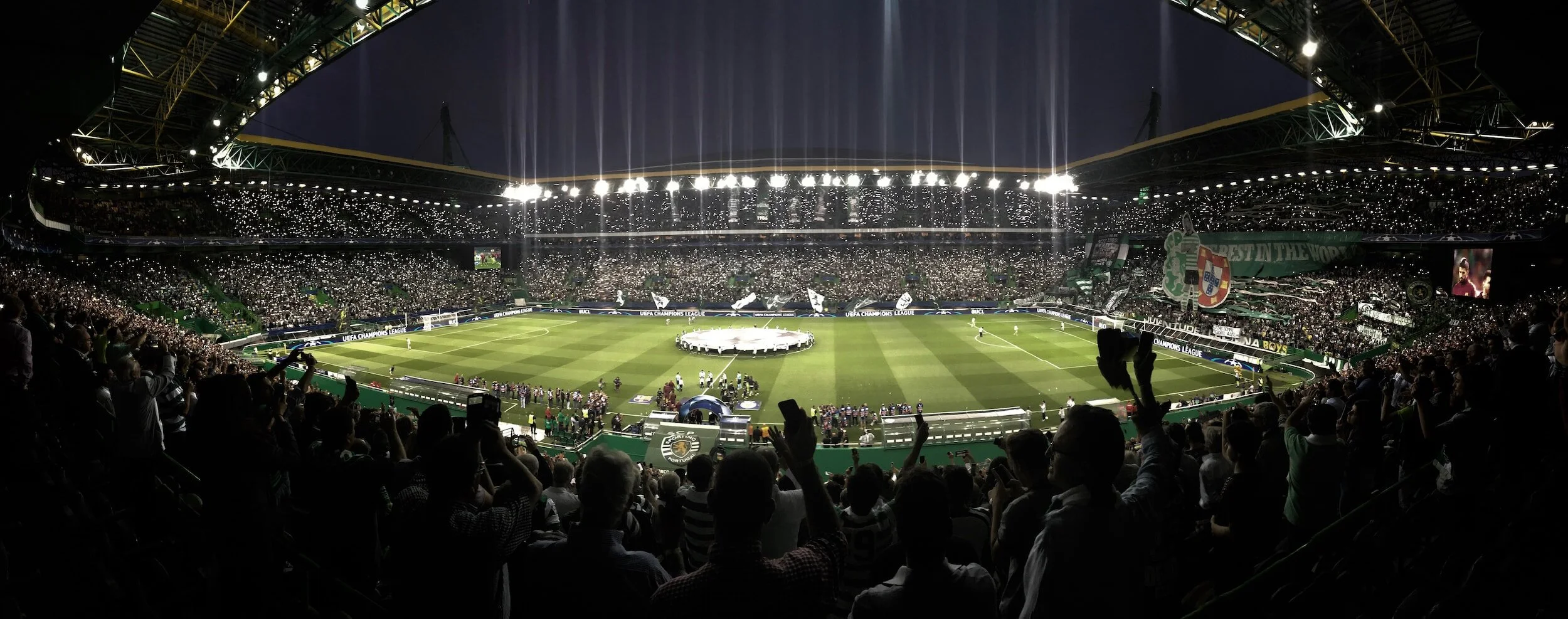 A packed soccer stadium filled with spectators, illuminated with bright stadium lights, preparing for a UEFA Champions League match.
