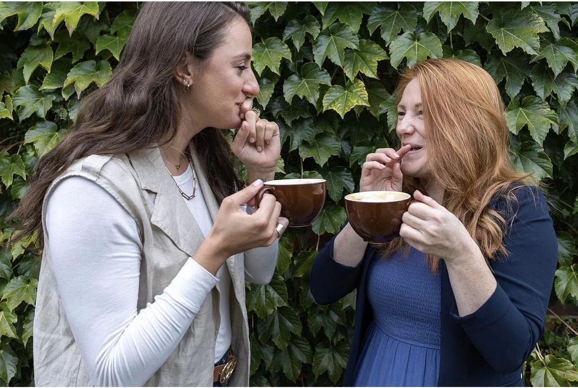 Two women sharing a moment, drinking coffee from brown cups, standing in front of green leafy plants.