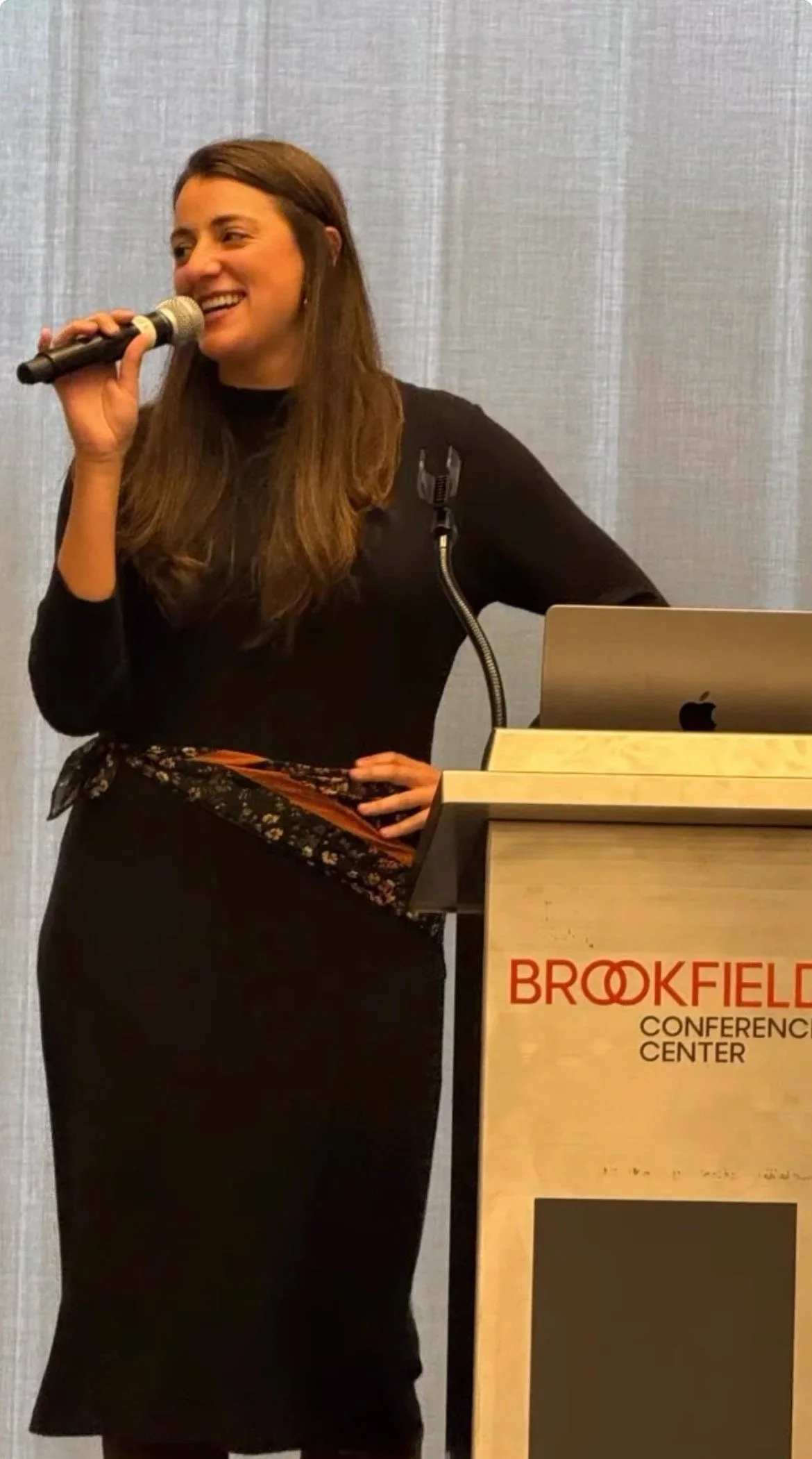 A woman is standing behind a podium at the Brookfield Conference Center, speaking into a microphone. She is wearing a black dress with a colorful scarf around her waist, and has long brown hair. A laptop is placed on the podium.