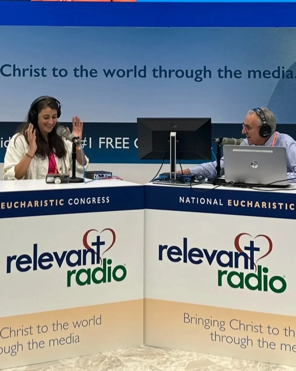 A radio broadcast studio with a woman smiling and a man speaking into microphones. The backdrop reads 'Christ to the world through the media,' and the front of the table features the logo 'relevant radio' with the slogan 'Bringing Christ to the world through the media.'