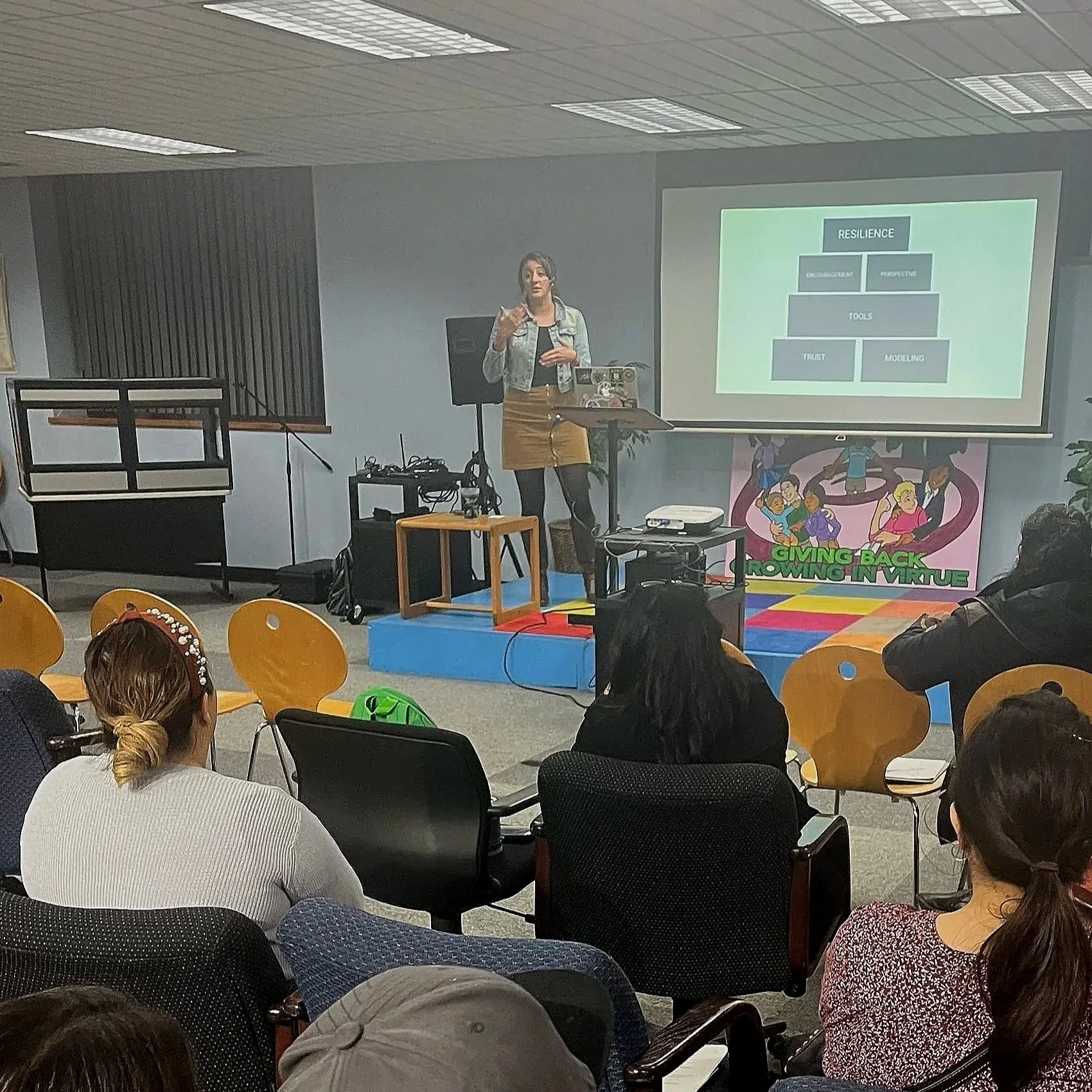 A woman giving a presentation in front of an audience, with a projection screen displaying a flowchart about resilience. The audience is seated, listening attentively.