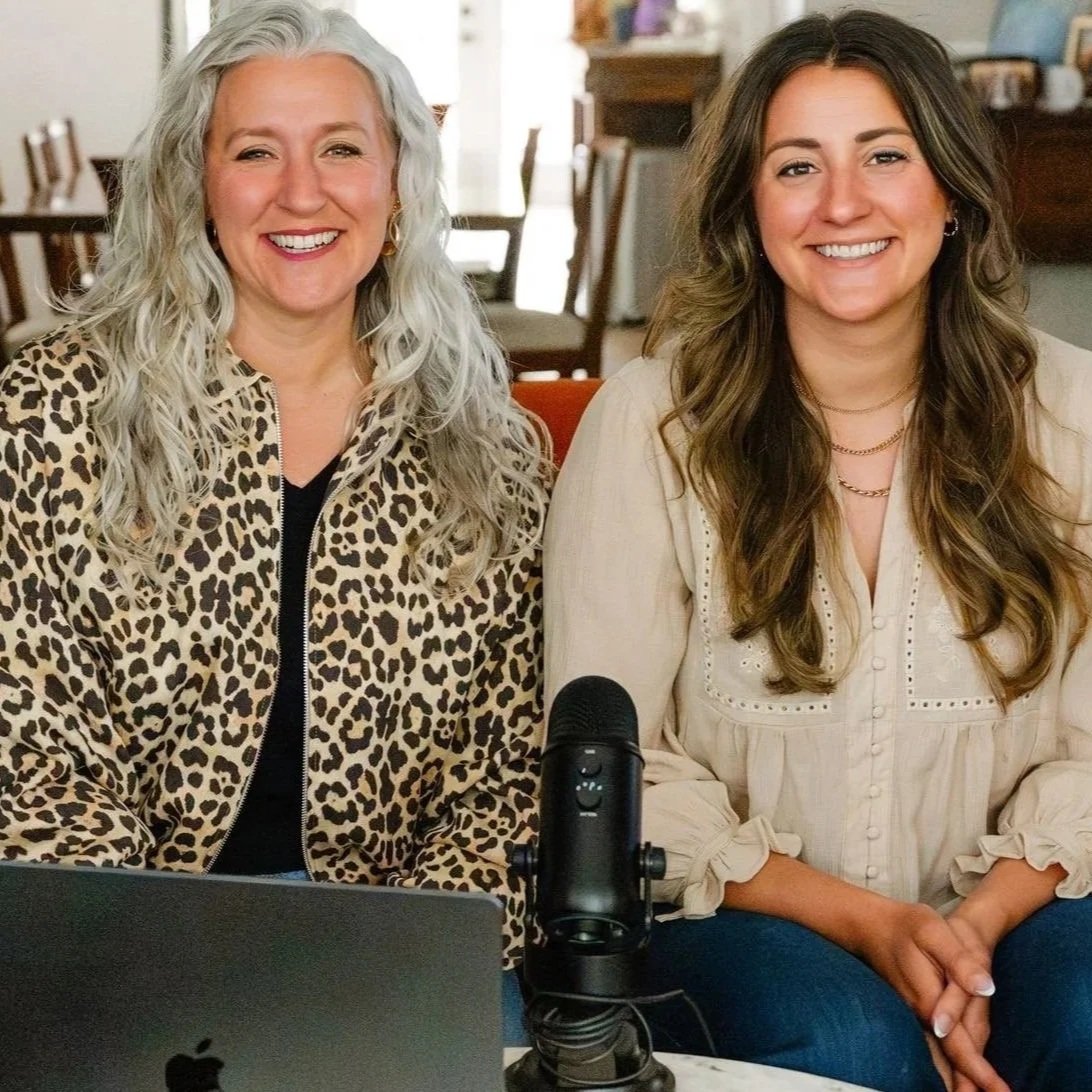 Two women sitting at a table smiling, with a microphone and a laptop in front of them, in a cozy indoor setting.